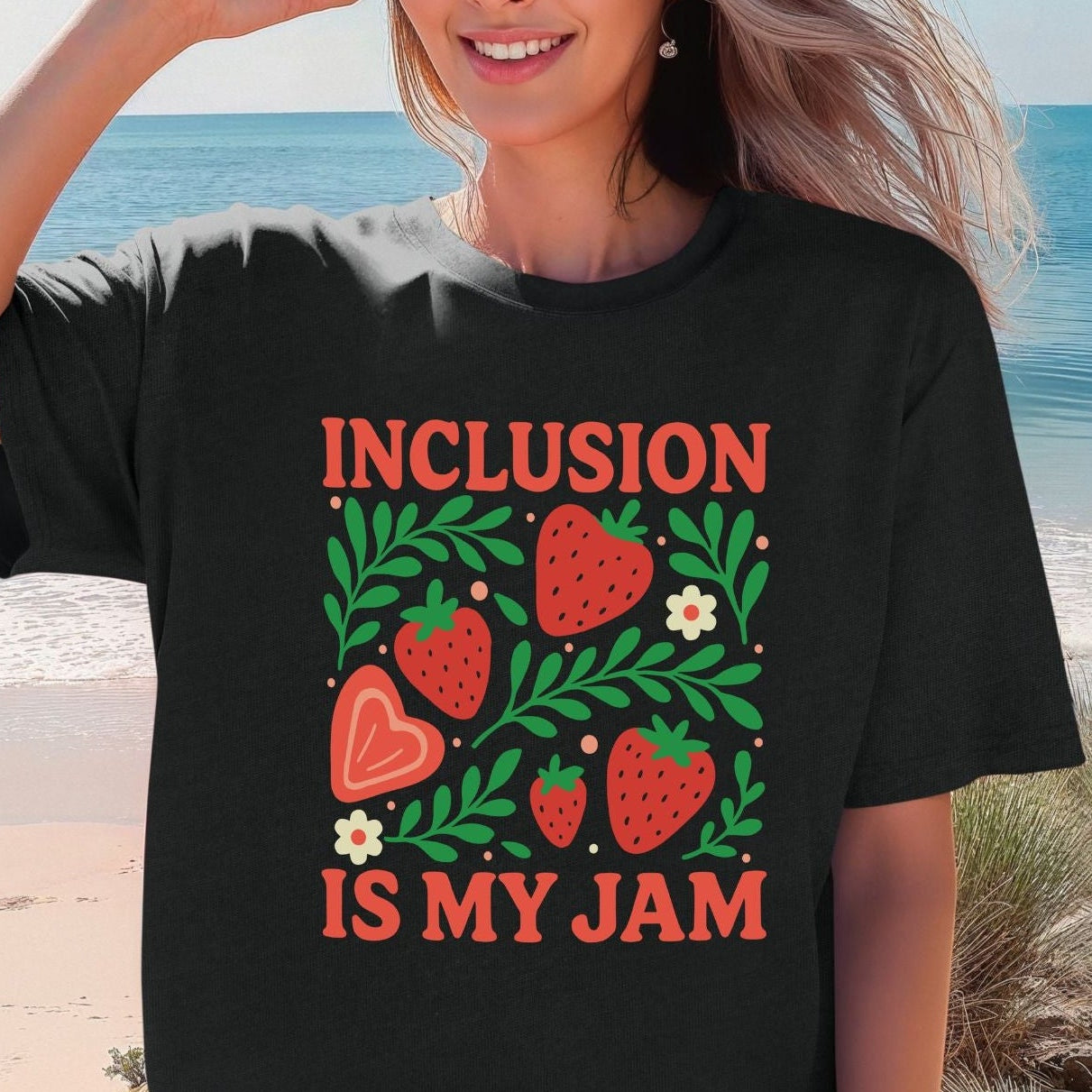 A smiling woman on a beach wearing a black oversized unisex t-shirt. The shirt features a vibrant botanical strawberry graphic with red strawberries in various sizes, a peach heart-shaped strawberry, lush green leafy branches, small white daisy flowers, and coral orange dot accents. The words 'Inclusion Is My Jam' are printed in bold coral red retro lettering above and below the graphic