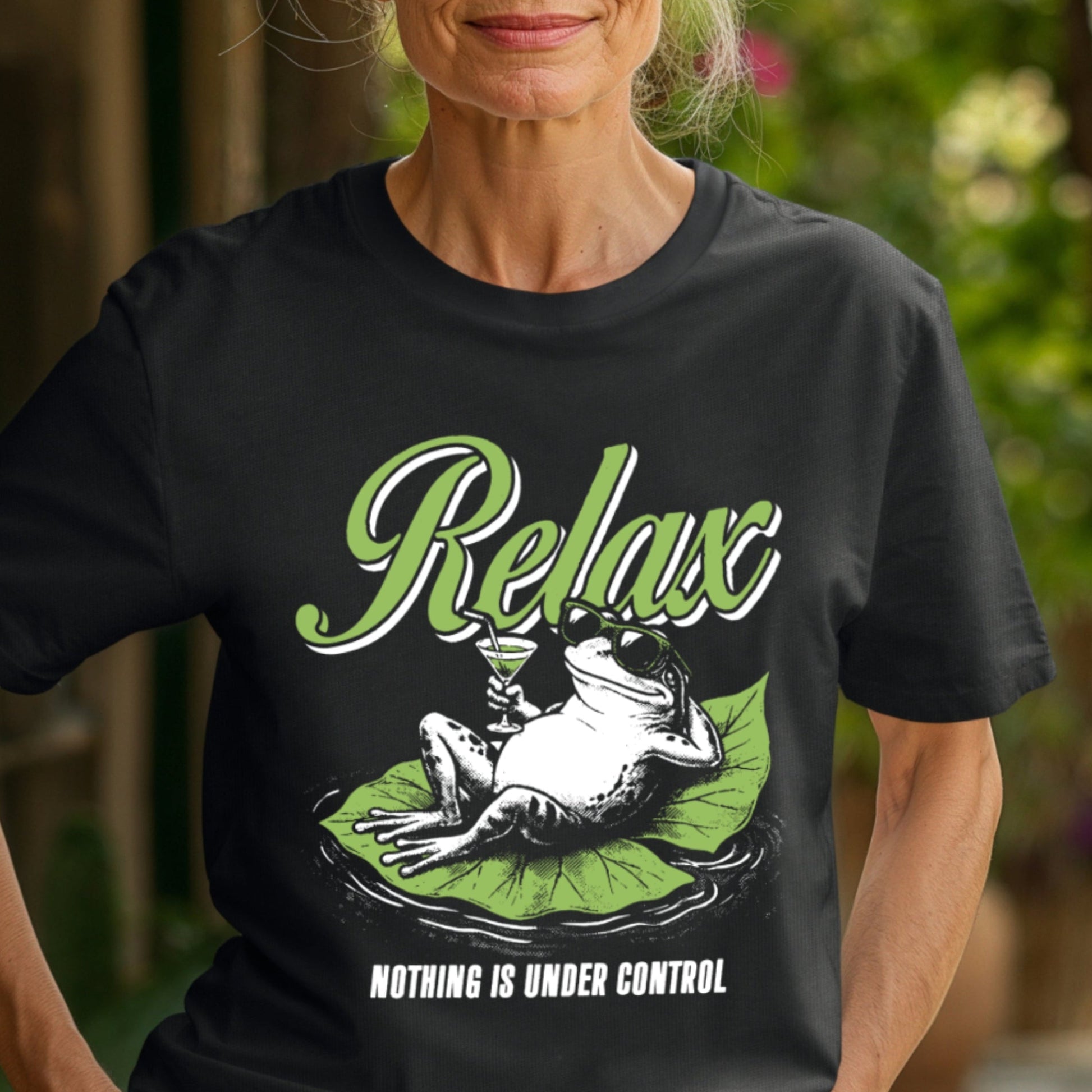 Older woman in a garden courtyard wearing a Black Comfort Colors unisex t-shirt. The shirt features a retro illustrated frog wearing sunglasses, lounging on a green lily pad while holding a cocktail glass. The word Relax appears in bold green script above and the phrase Nothing Is Under Control is printed in white block letters below.
