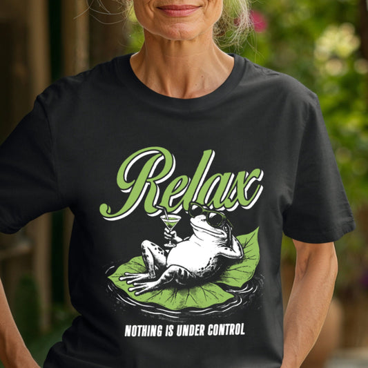 Older woman in a garden courtyard wearing a Black Comfort Colors unisex t-shirt. The shirt features a retro illustrated frog wearing sunglasses, lounging on a green lily pad while holding a cocktail glass. The word Relax appears in bold green script above and the phrase Nothing Is Under Control is printed in white block letters below.