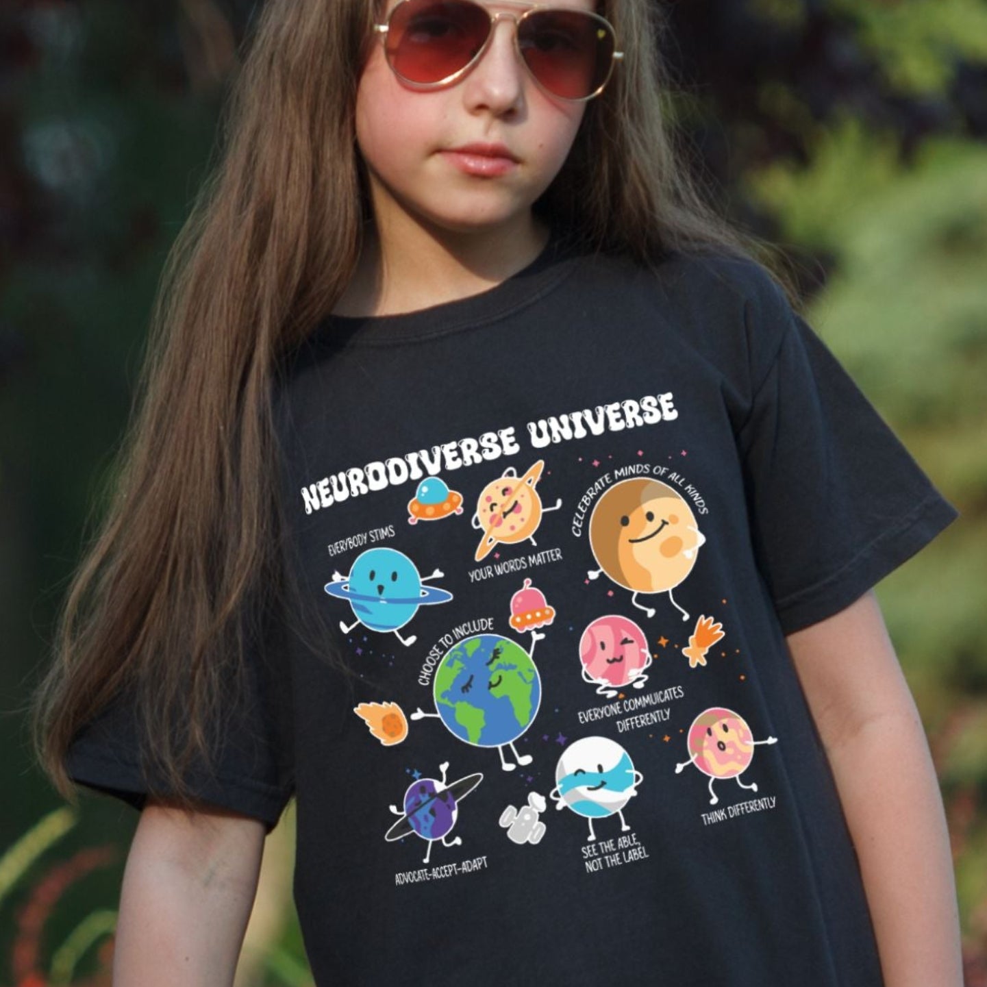 Young girl outdoors wearing a Black Comfort Colors youth t-shirt. The shirt features colorful cartoon planet characters each labeled with an empowering neurodiversity message including Everybody Stims, Your Words Matter, Choose to Include, and Think Differently. The title Neurodiverse Universe is printed at the top in bold white lettering.