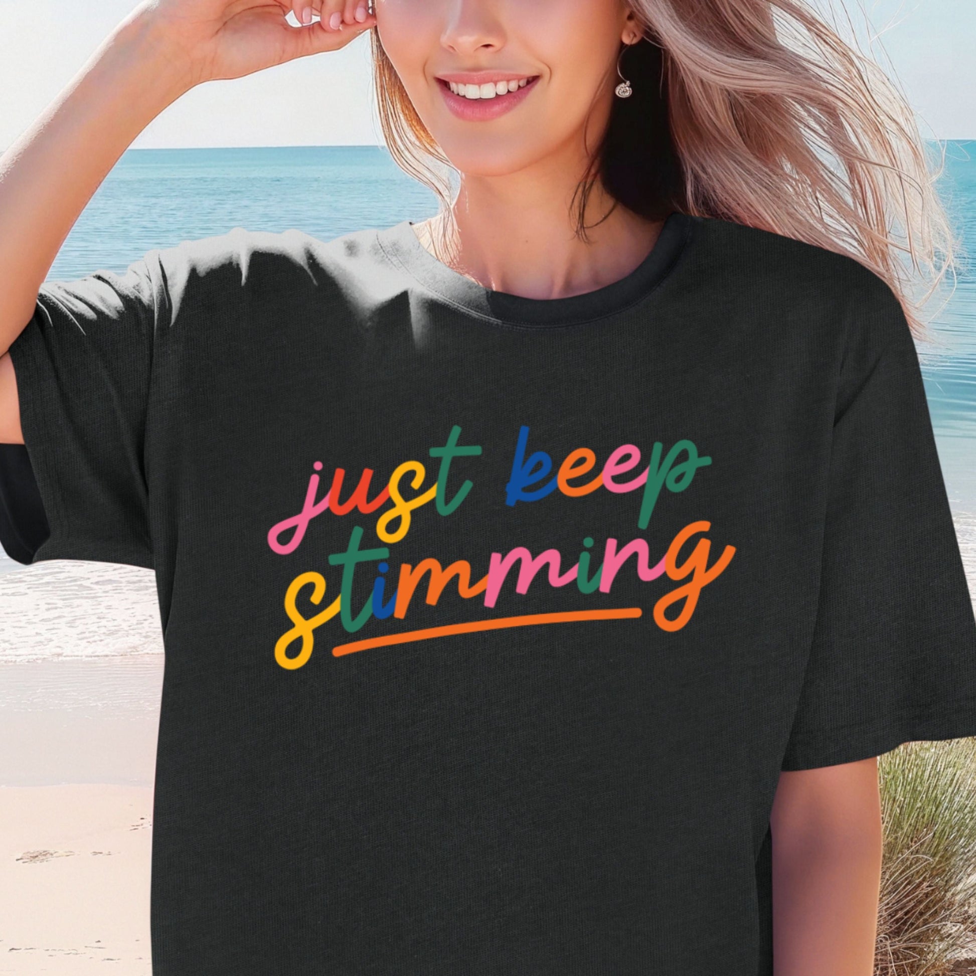 Smiling woman on a beach wearing a black oversized unisex t-shirt. The shirt features the phrase just keep stimming in large multicolor rainbow script lettering in pink, blue, green, yellow, and orange with an orange underline beneath the word stimming.