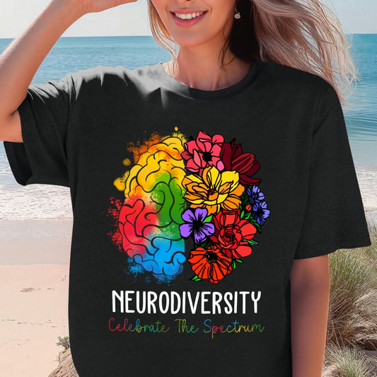 Woman on a beach wearing a black oversized t-shirt with a rainbow floral brain graphic. The left brain hemisphere is filled with orange, yellow, green, red, and blue watercolor splashes. The right side blooms into pink, red, yellow, purple, and orange wildflowers. Text reads NEURODIVERSITY and Celebrate The Spectrum in multicolor lettering.