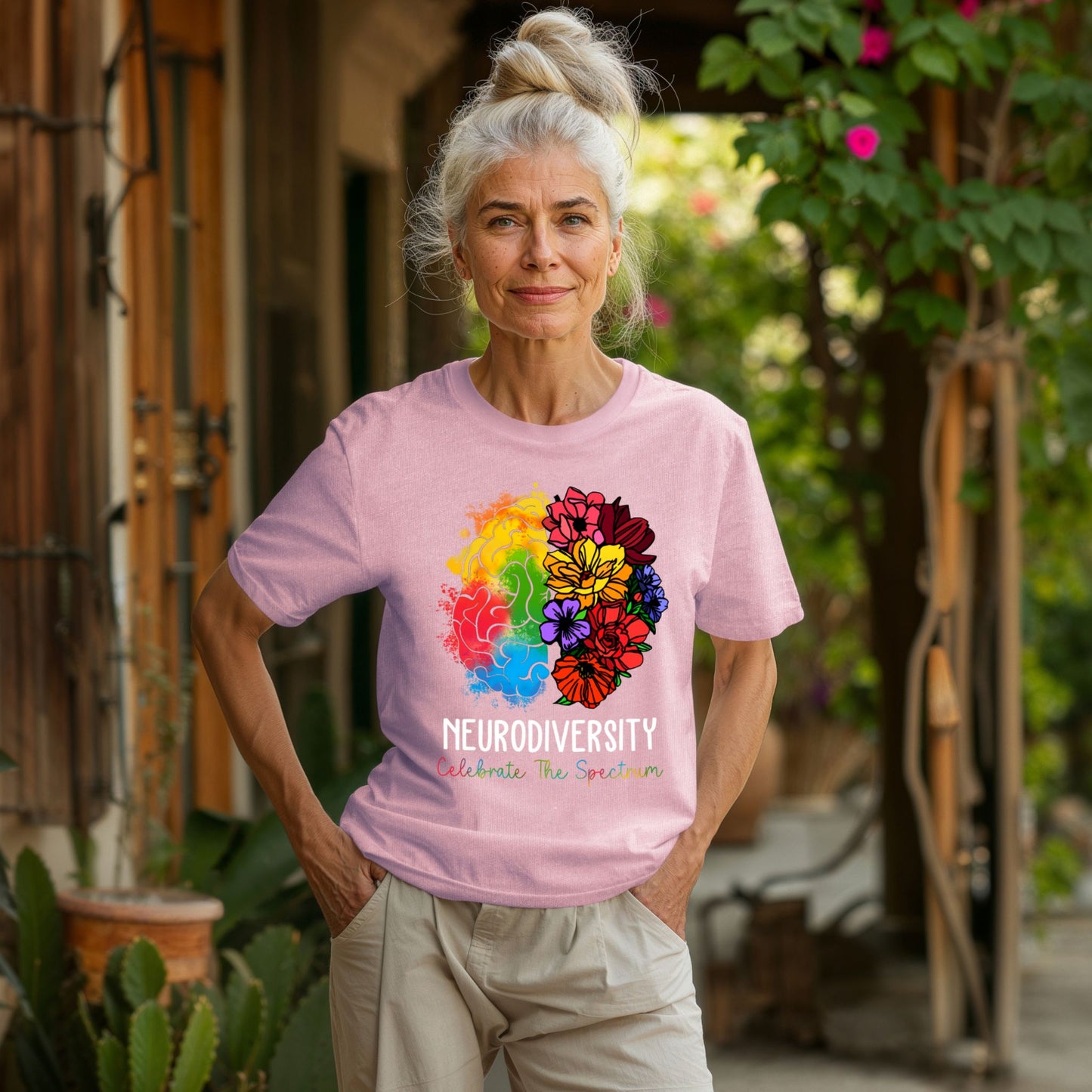 Older woman outdoors wearing a soft pink t-shirt with a rainbow floral brain graphic. The left brain side features colorful watercolor splashes and the right side blooms into pink, red, yellow, purple, and orange wildflowers. Text reads NEURODIVERSITY and Celebrate The Spectrum in multicolor lettering.