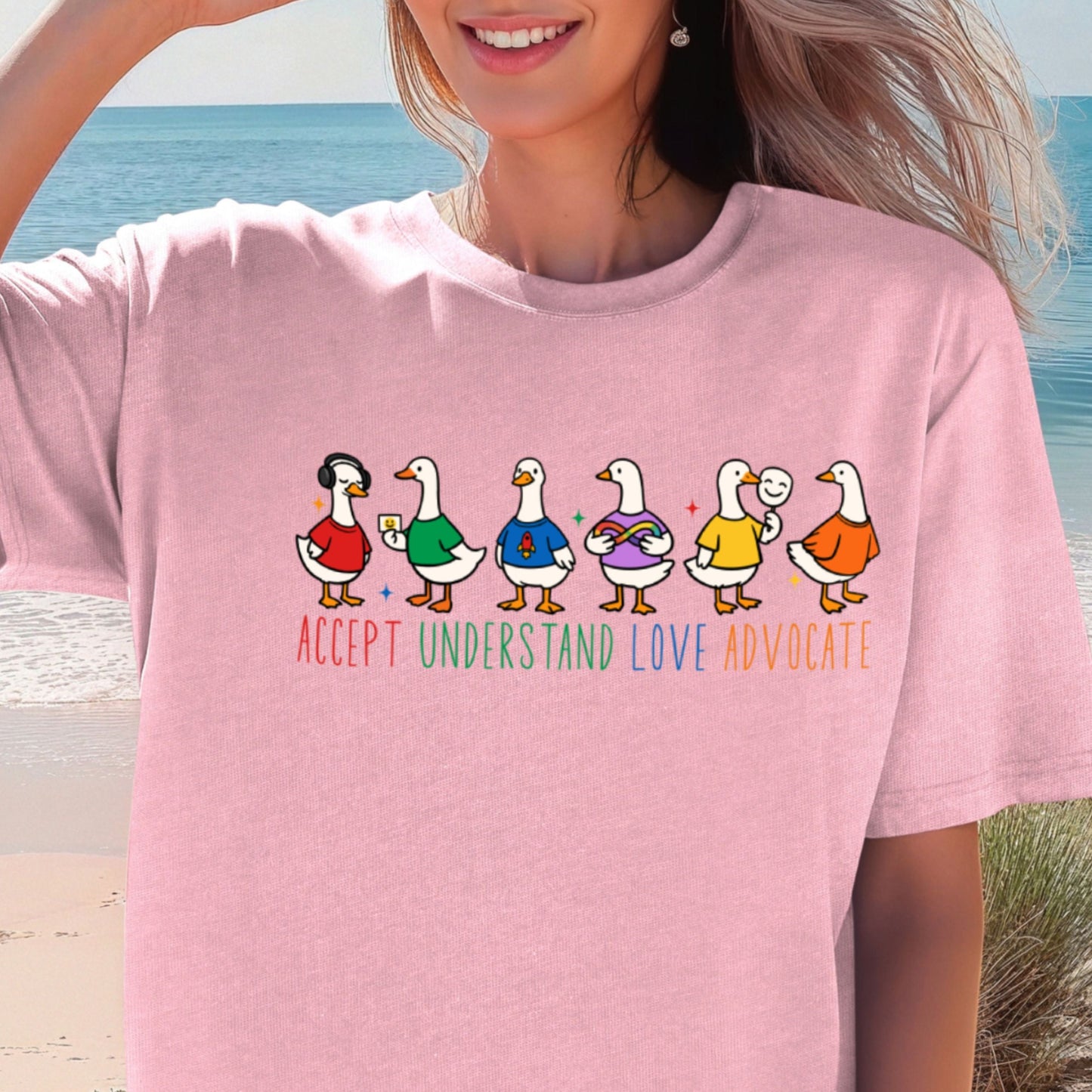 A smiling woman on a beach wearing a soft pink oversized unisex t-shirt. The shirt features six cartoon duck characters lined up in a row, each wearing a different colorful outfit in red, green, blue, purple, yellow, and orange. Below the ducks are the words Accept Understand Love Advocate printed in multicolor lettering with sparkle accents.