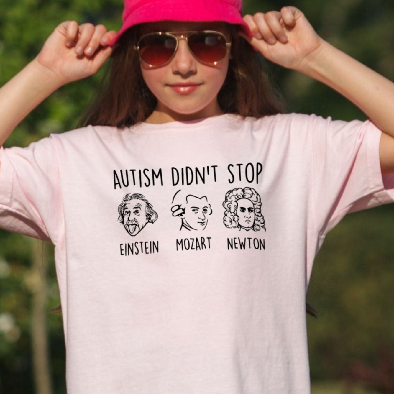 Young girl outdoors wearing a Blossom soft pink Comfort Colors youth t-shirt. The shirt features hand-drawn style portraits of Einstein, Mozart, and Newton in black ink with the text Autism Didn't Stop printed above in bold black lettering and each name labeled below their portrait.