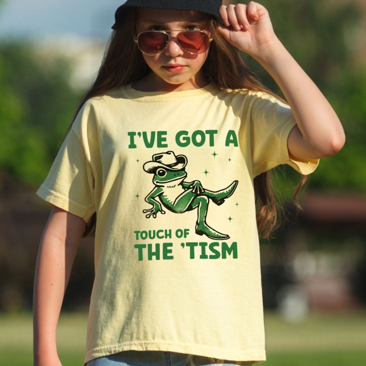 Young girl outdoors wearing a Butter pale yellow Comfort Colors youth t-shirt. The shirt features a green illustrated frog wearing a cowboy hat and cowboy boots with sparkle accents. Text above reads I've Got a and text below reads Touch of The Tism in bold green lettering.
