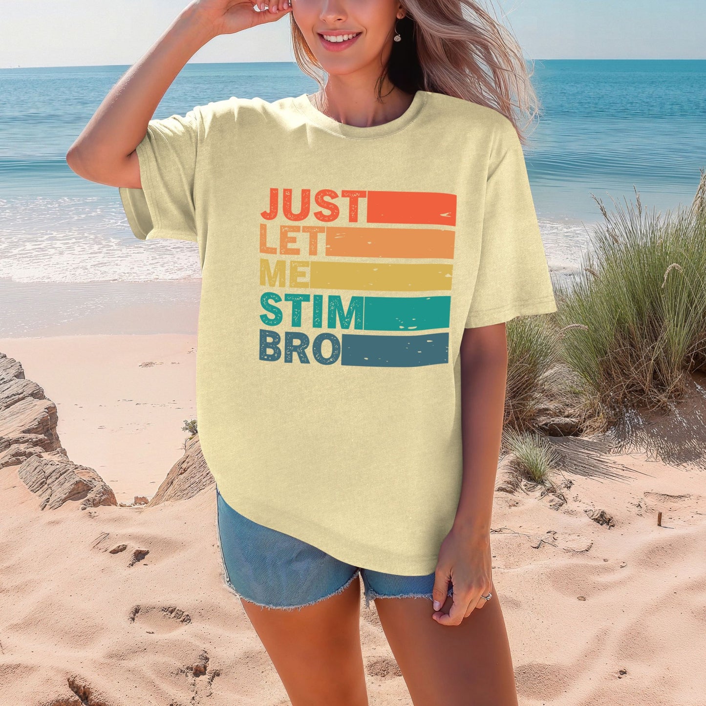 Smiling woman on a beach wearing a Butter pale yellow Comfort Colors oversized t-shirt. The shirt features bold retro stripe lettering reading Just Let Me Stim Bro stacked vertically with each word on a colored stripe in orange, peach, yellow, teal, and dark teal with a distressed vintage texture.