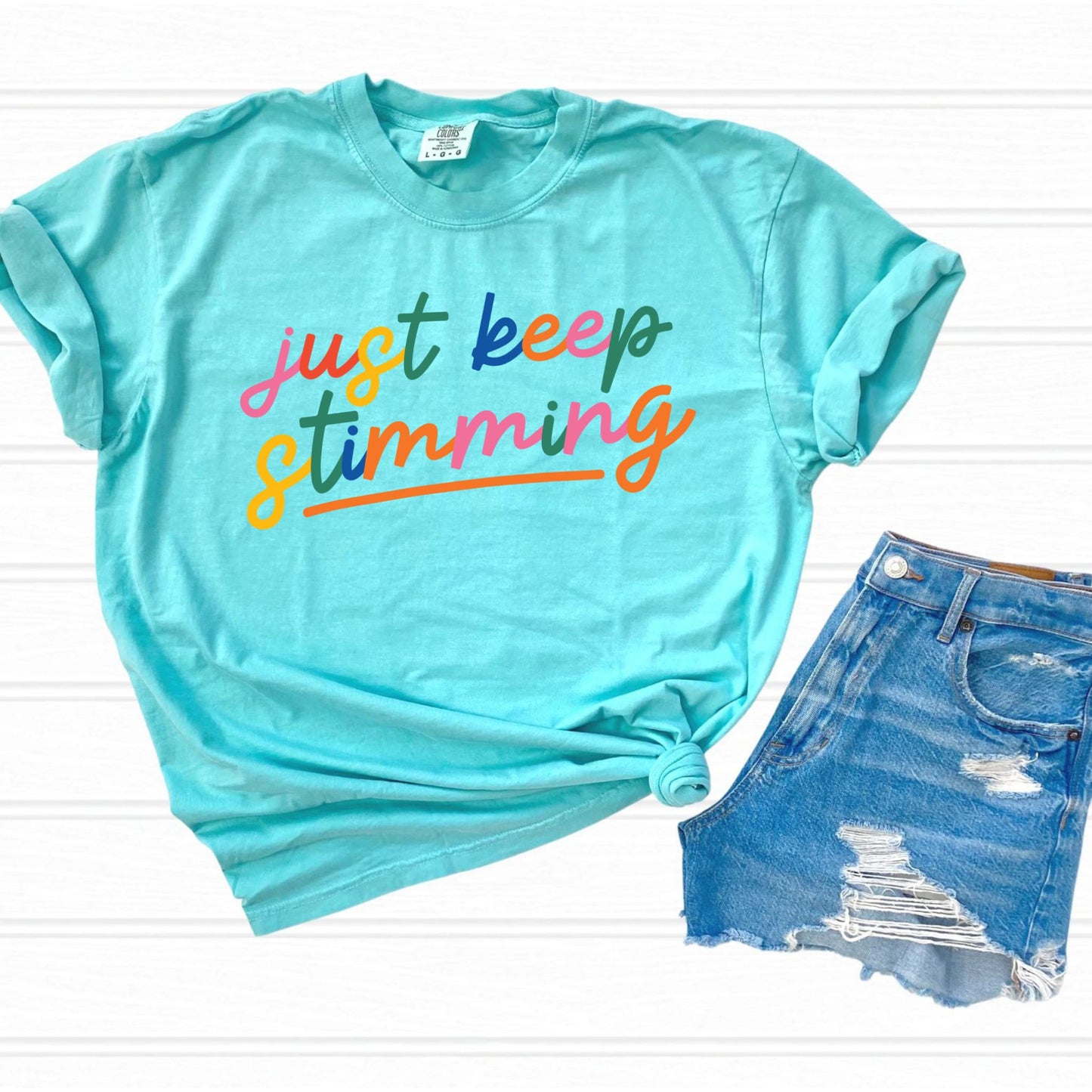Flat lay of a chalky mint green t-shirt with rolled sleeves next to a pair of denim shorts. The shirt features the phrase just keep stimming in large multicolor rainbow script lettering in pink, blue, green, yellow, and orange with an orange underline beneath the word stimming.