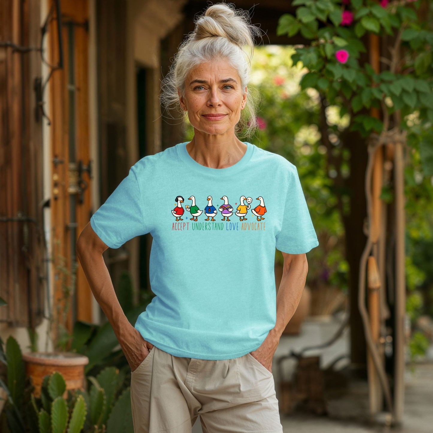 An older woman with white hair standing outdoors wearing a light mint green unisex t-shirt. The shirt displays six cheerful cartoon duck characters in a row wearing colorful outfits. The text below reads Accept Understand Love Advocate in rainbow colored lettering.