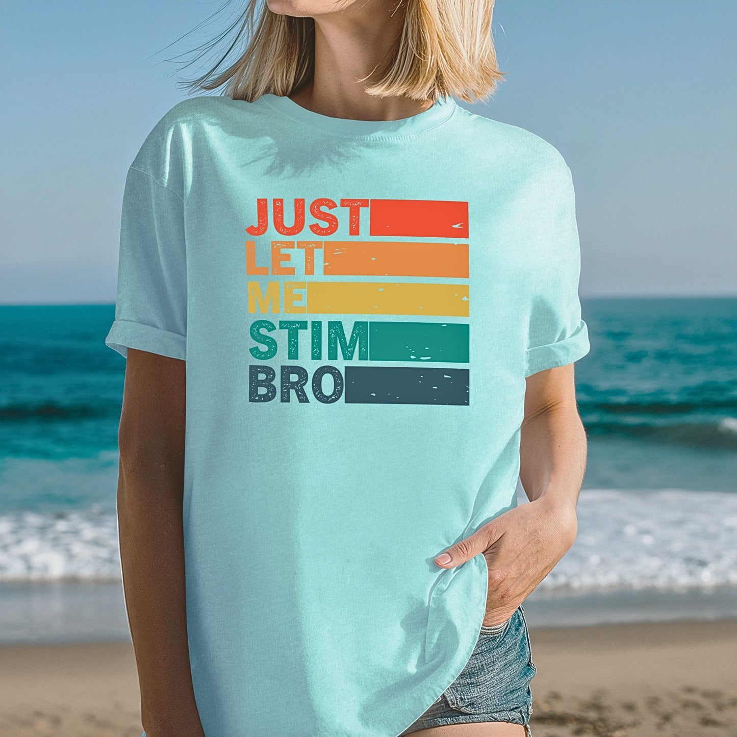 Woman at the beach wearing a Chalky Mint light mint green Comfort Colors t-shirt with rolled sleeves. The shirt features bold retro stripe lettering reading Just Let Me Stim Bro stacked vertically with each word on a colored stripe in orange, peach, yellow, teal, and dark teal with a distressed vintage texture.