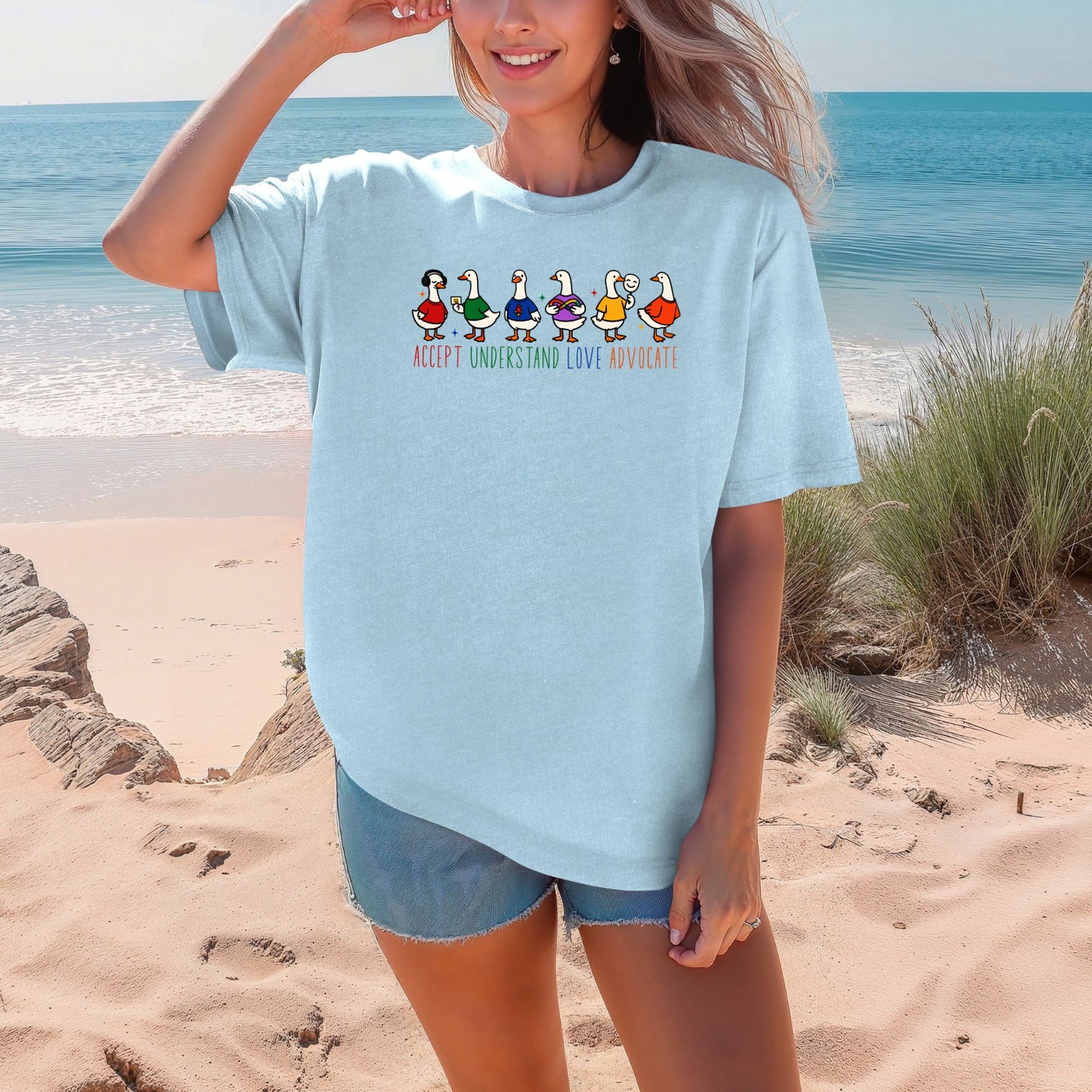 A smiling woman on a beach wearing a light blue chambray oversized unisex t-shirt. The shirt features six adorable cartoon ducks wearing colorful outfits in red, green, blue, purple, yellow, and orange. The words Accept Understand Love Advocate are printed below in multicolor lettering with sparkle accents.