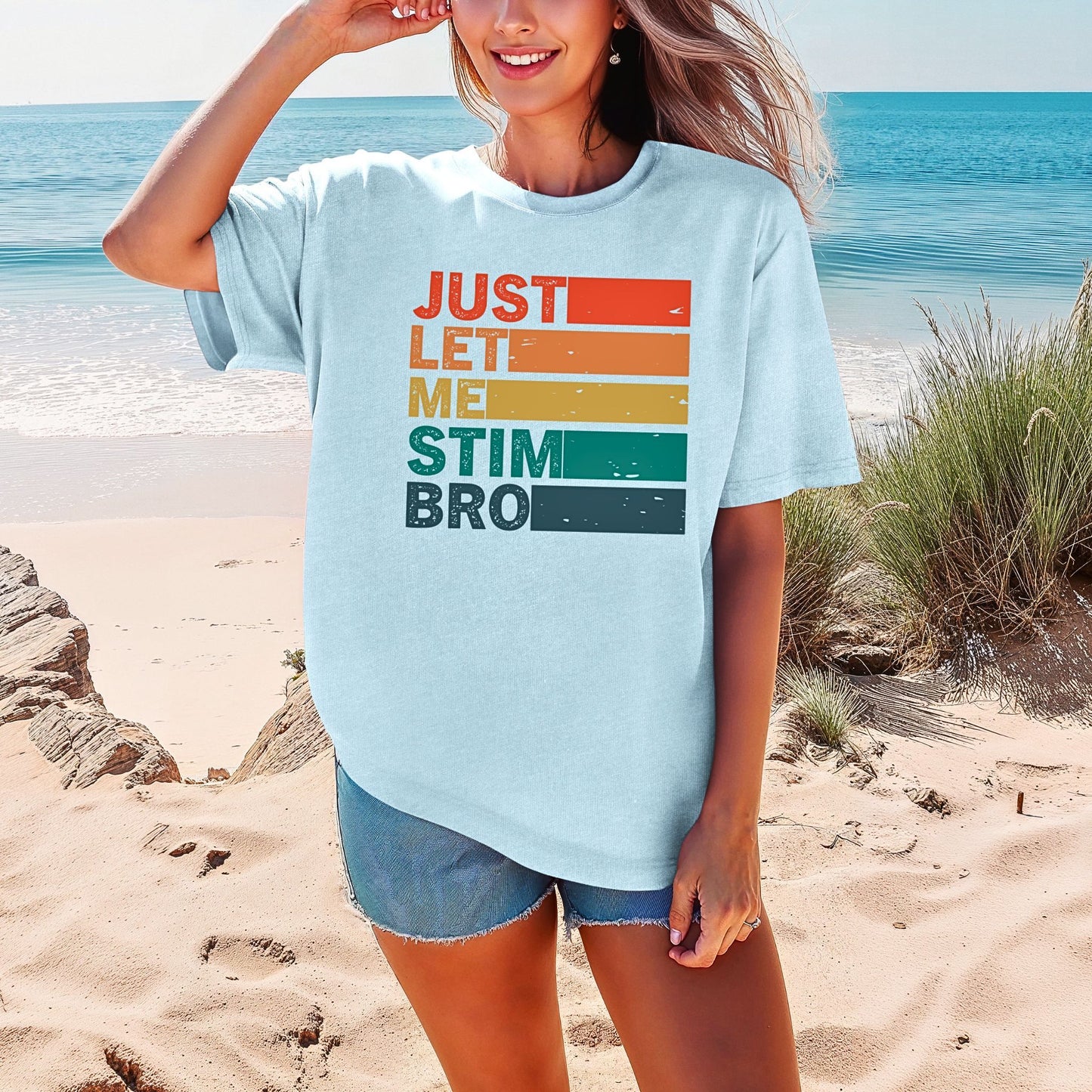 Smiling woman on a beach wearing a Chambray light blue Comfort Colors oversized t-shirt. The shirt features bold retro stripe lettering reading Just Let Me Stim Bro stacked vertically with each word on a colored stripe in orange, peach, yellow, teal, and dark teal with a distressed vintage texture.