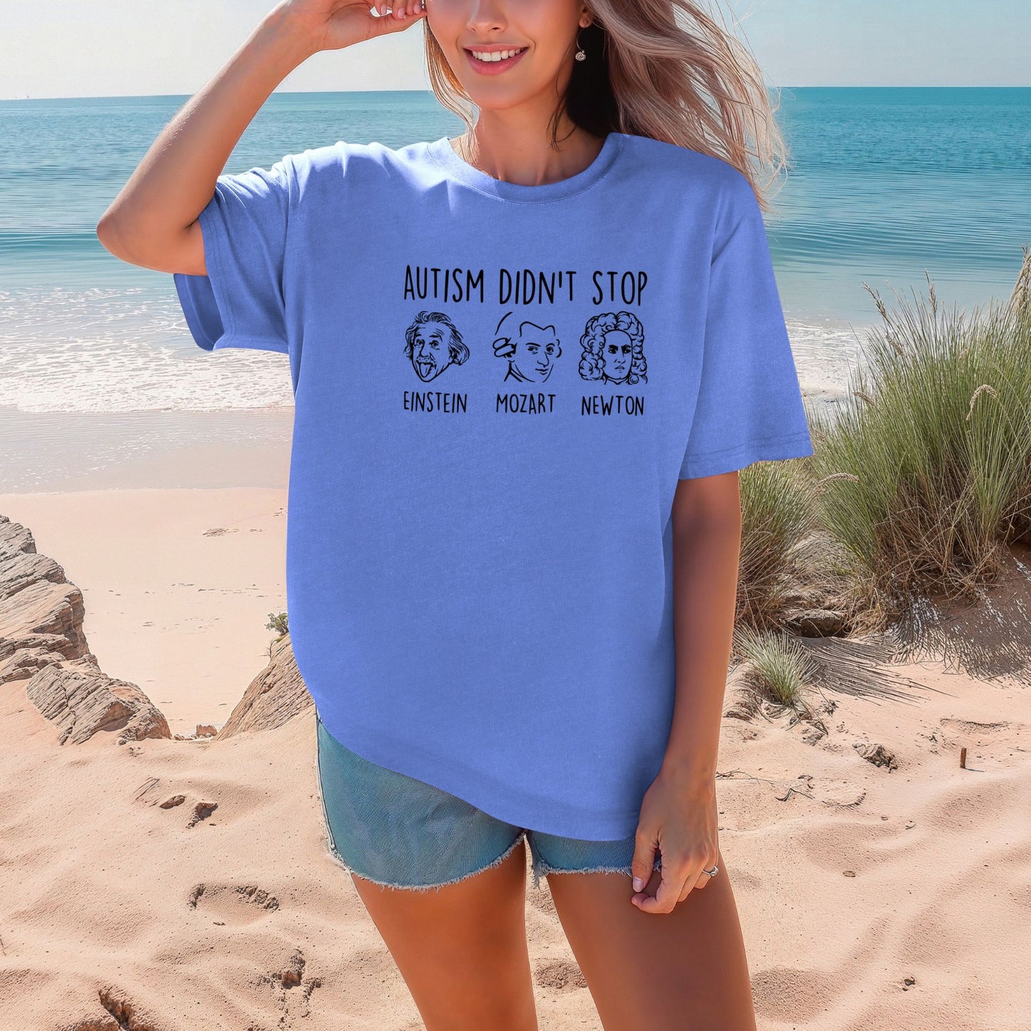 Smiling woman on a beach wearing a Flo Blue periwinkle blue Comfort Colors oversized t-shirt. The shirt features hand-drawn style portraits of Einstein, Mozart, and Newton in black ink with the text Autism Didn't Stop printed above in bold black lettering and each name labeled below their portrait.