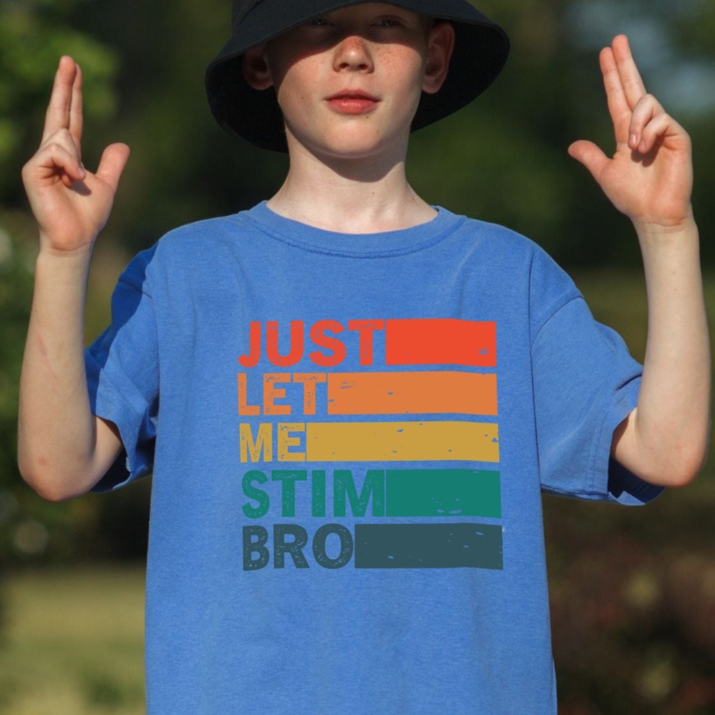 Young boy outdoors wearing a Flo Blue bright blue Comfort Colors youth t-shirt. The shirt features bold retro stripe lettering reading Just Let Me Stim Bro stacked vertically with each word on a colored stripe in orange, peach, yellow, green, and teal with a distressed vintage texture.