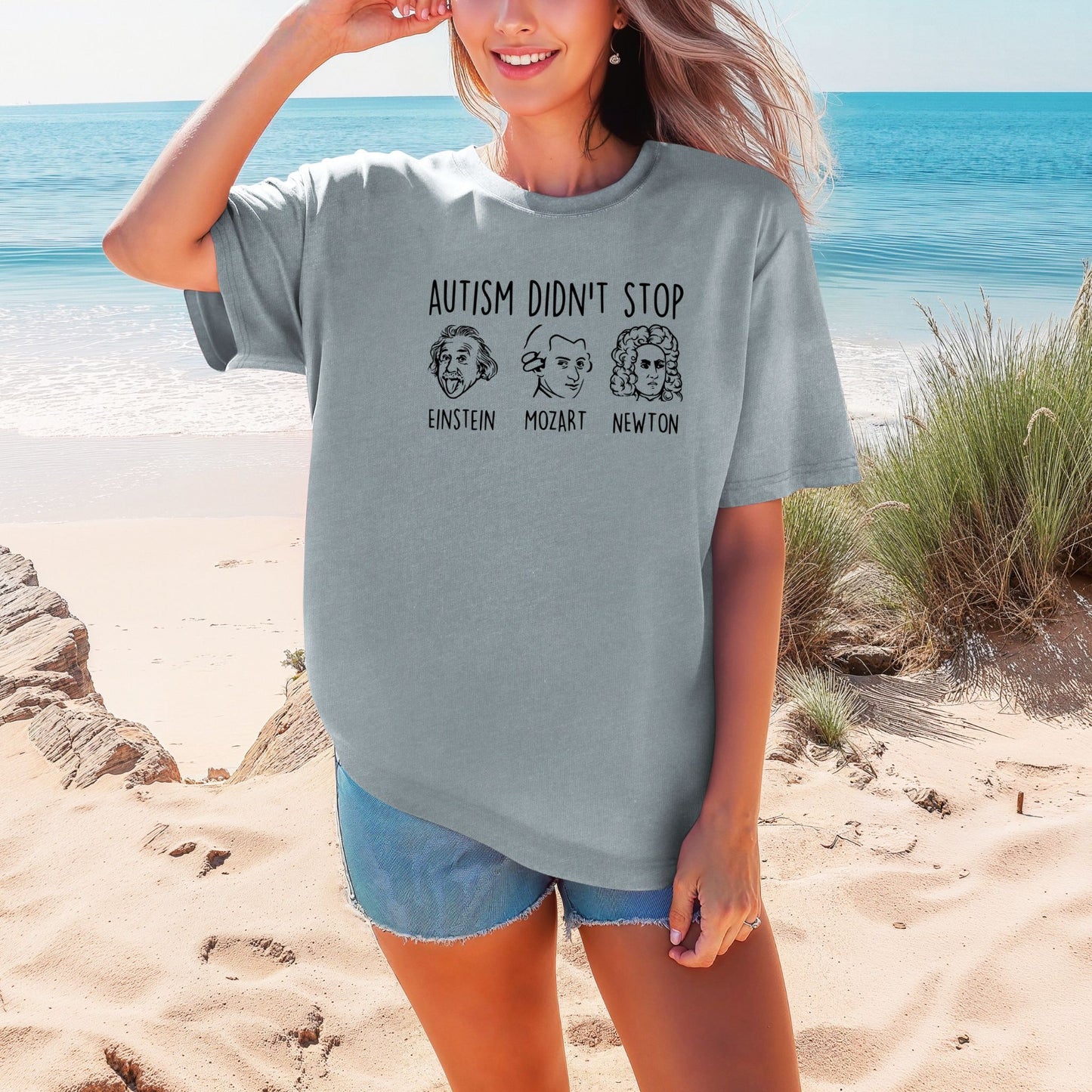 Smiling woman on a beach wearing a Granite heathered blue-gray Comfort Colors oversized t-shirt. The shirt features hand-drawn style portraits of Einstein, Mozart, and Newton in black ink with the text Autism Didn't Stop printed above in bold black lettering and each name labeled below their portrait.