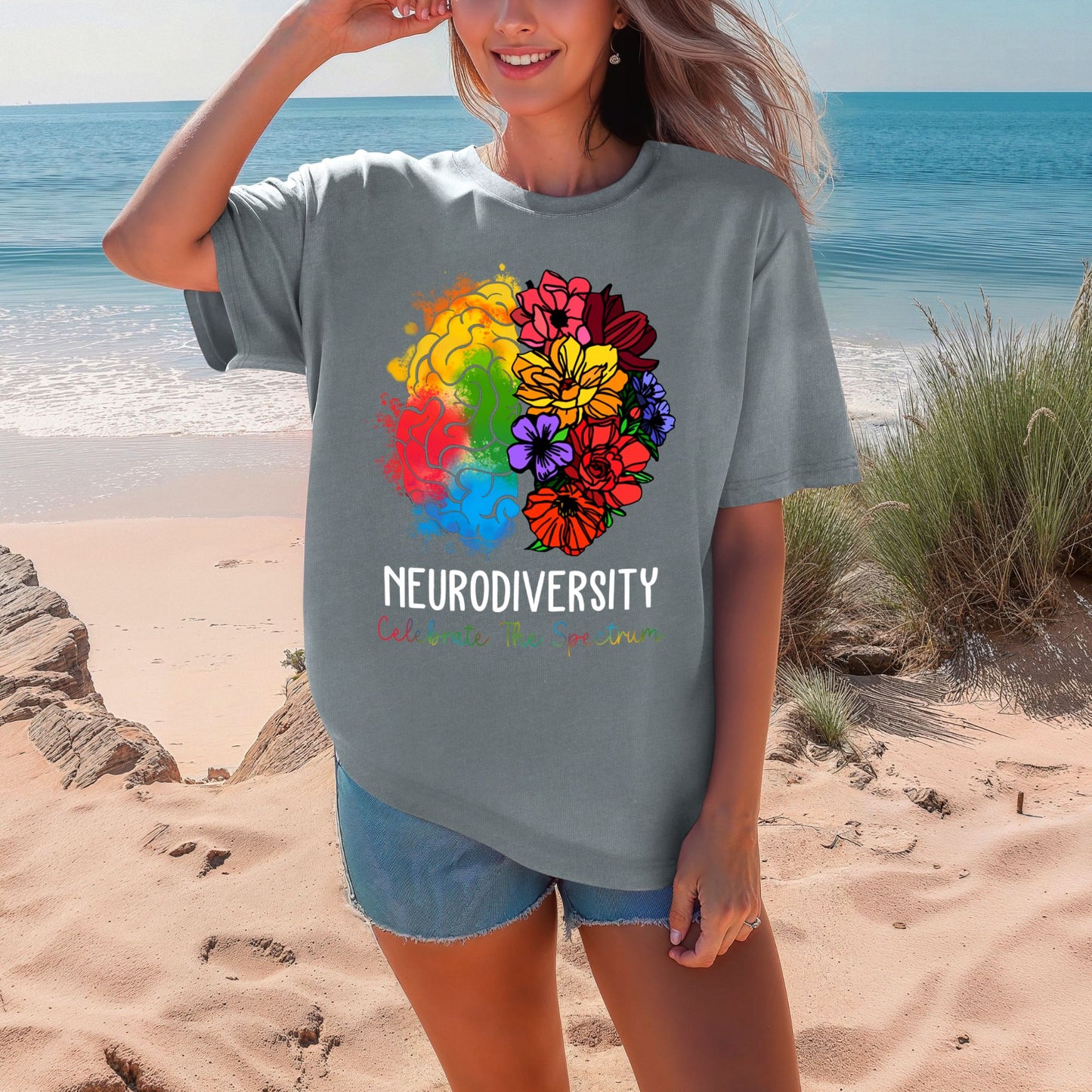 Woman on a beach wearing a heathered blue-gray oversized t-shirt with a vibrant rainbow floral brain graphic. The left brain has orange, yellow, green, red, and blue watercolor splashes. The right side bursts into colorful wildflowers. Text reads NEURODIVERSITY and Celebrate The Spectrum in multicolor lettering.