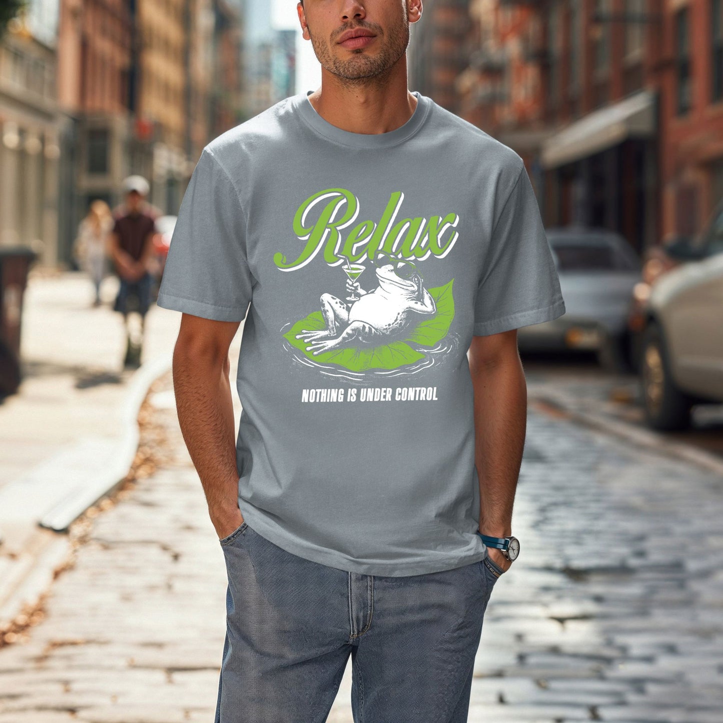 Man on a city street wearing a Granite heathered blue-gray Comfort Colors unisex t-shirt. The shirt features a retro illustrated frog wearing sunglasses, lounging on a green lily pad holding a cocktail glass. The word Relax appears in bold green script above and Nothing Is Under Control in white block letters below.