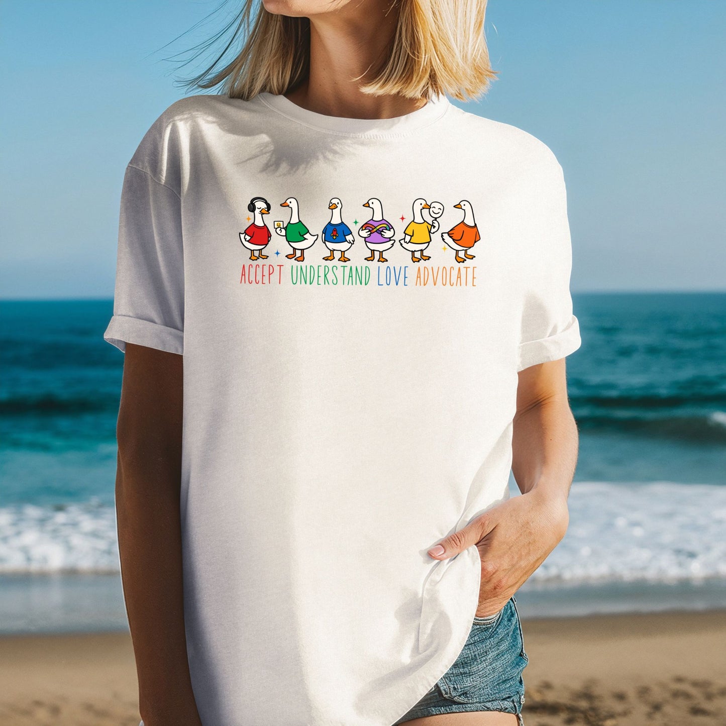 A woman at the beach wearing a cream ivory unisex t-shirt with rolled sleeves. The shirt features a row of six cartoon duck characters each wearing a different colorful outfit. Below the ducks the text reads Accept Understand Love Advocate in bright multicolor lettering.