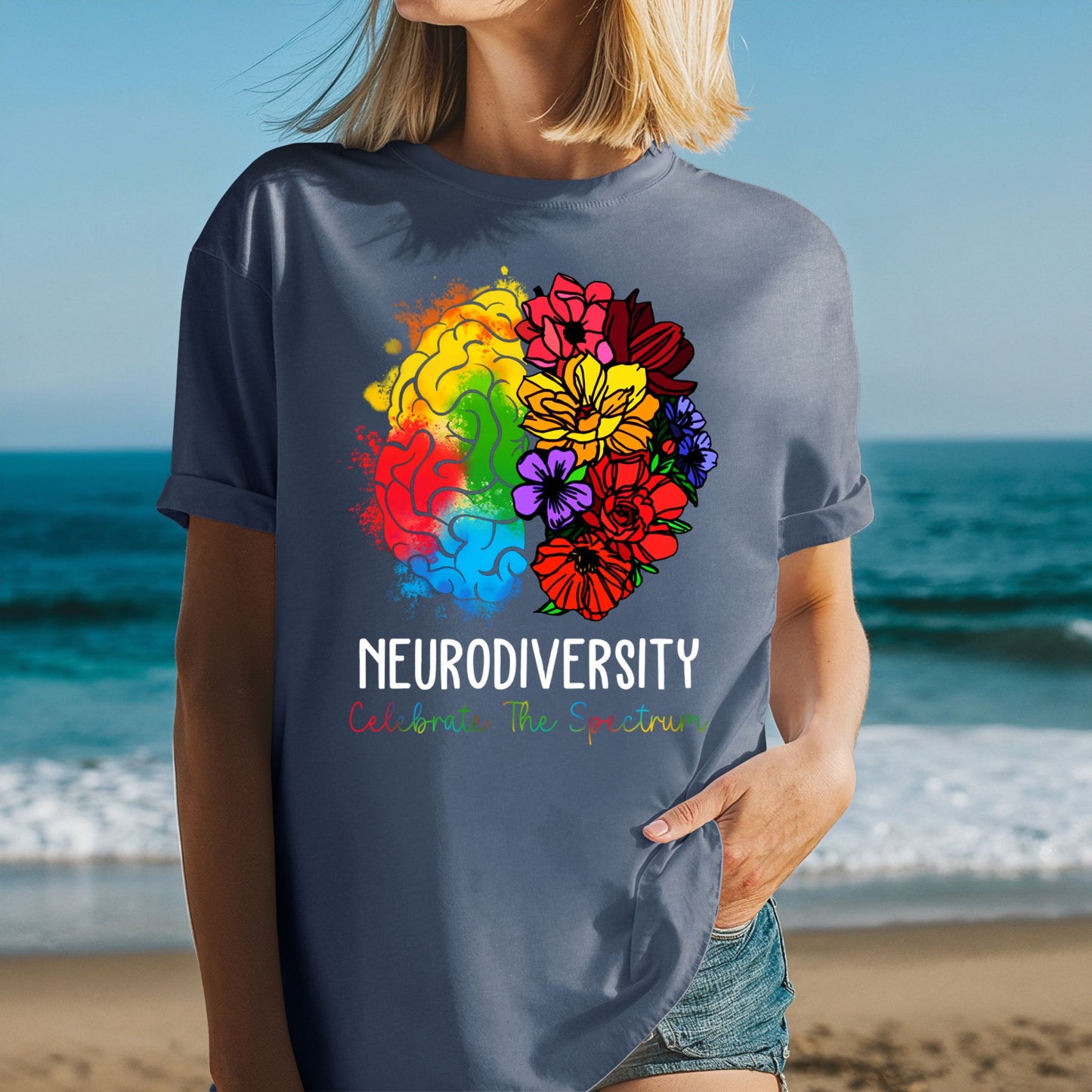 Woman at the beach wearing a medium blue jean-toned t-shirt with rolled sleeves and a bold rainbow floral brain graphic. The left brain is painted in rainbow watercolor and the right side bursts into pink, red, yellow, purple, and orange wildflowers. Text reads NEURODIVERSITY and Celebrate The Spectrum in multicolor lettering.