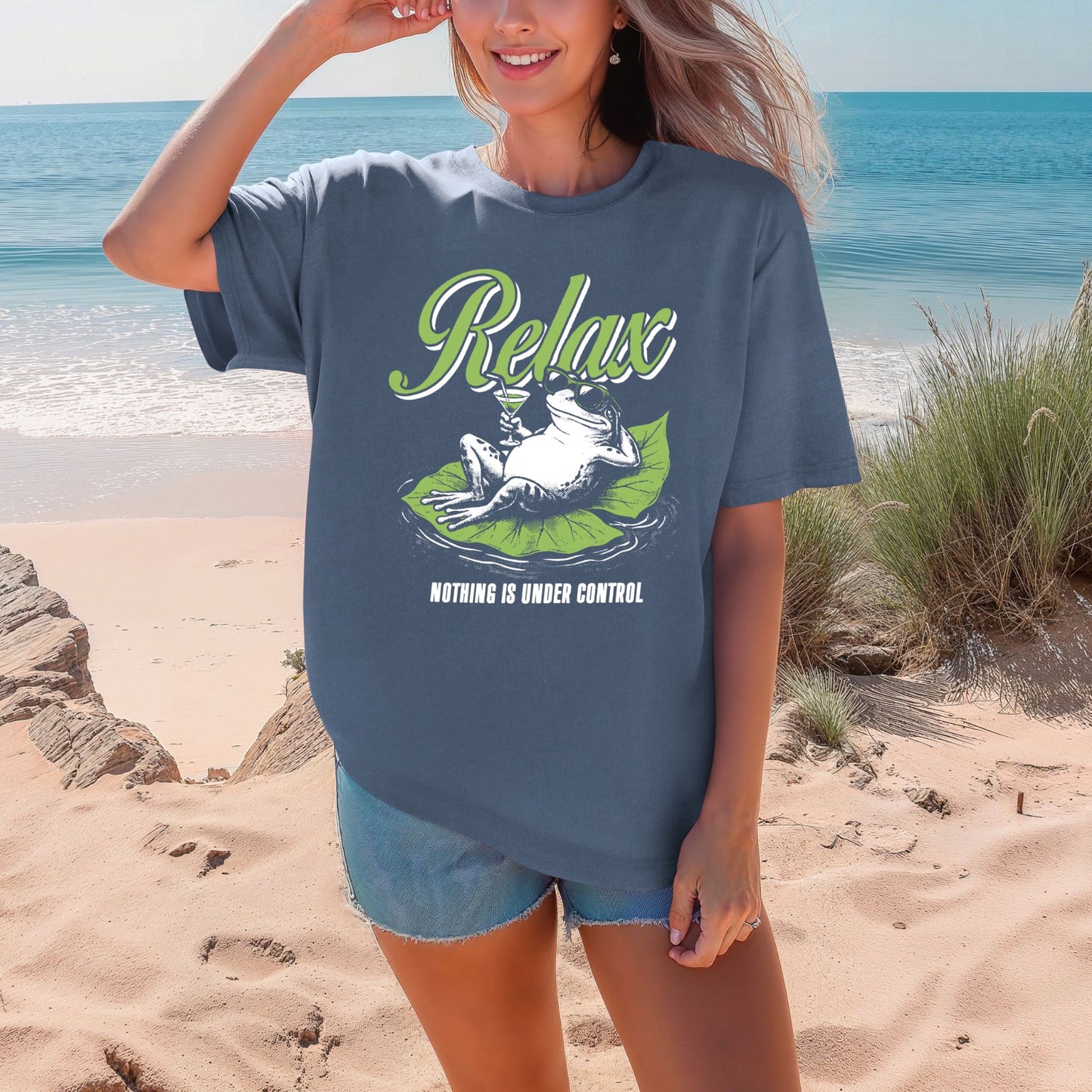 Smiling woman on a beach wearing a Jean Blue medium blue Comfort Colors oversized unisex t-shirt. The shirt features a retro illustrated frog wearing sunglasses, lounging on a green lily pad holding a cocktail glass. The word Relax appears in bold green script above and Nothing Is Under Control in white block letters below.
