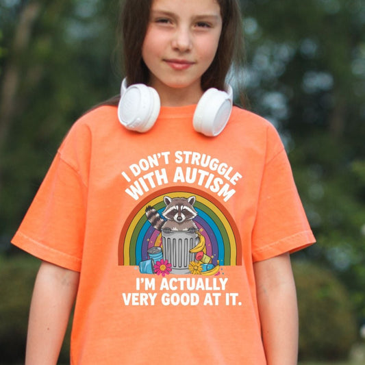 Comfort Colors 9018 youth t-shirt in Melon orange. A young girl stands in a grassy park holding a stuffed animal with white headphones around her neck. The shirt features a raccoon in a trash can in front of a rainbow with the text I Don't Struggle With Autism I'm Actually Very Good At It.