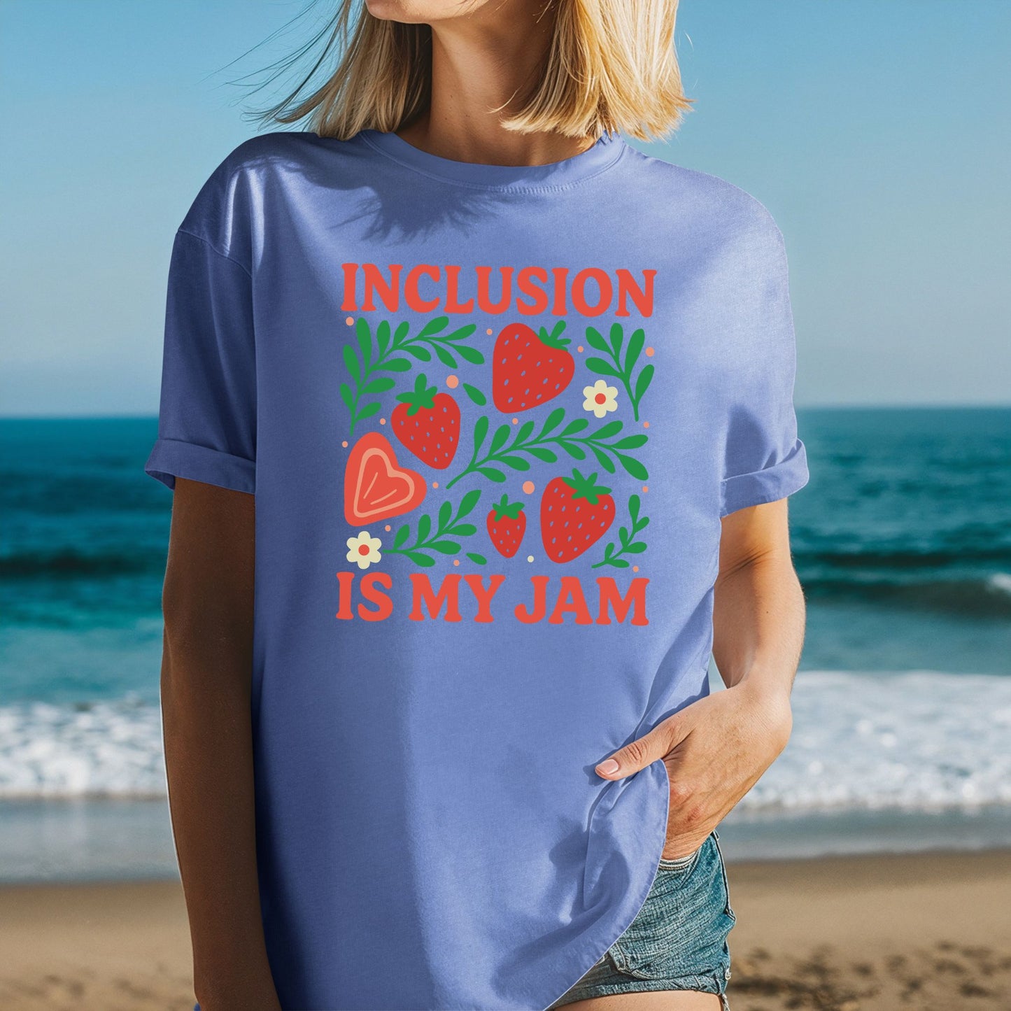 A woman at the beach wearing a Mystic Blue oversized unisex t-shirt with rolled sleeves. The shirt features a bold strawberry garden graphic with five red strawberries of varying sizes, a large peach heart-shaped strawberry, lush green leafy branches spreading throughout, small cream daisy flowers with orange centers, and scattered peach dot accents. The words 'Inclusion Is My Jam' are printed in bold coral red retro lettering above and below the graphic.