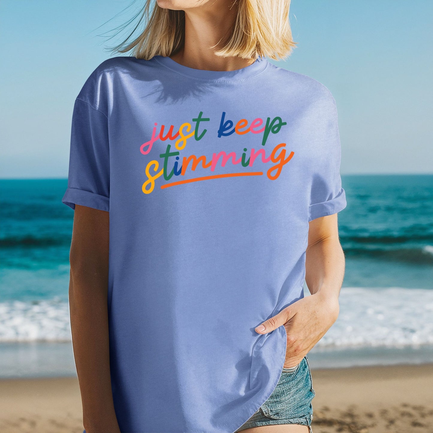 Woman at the beach wearing a periwinkle mystic blue t-shirt with rolled sleeves. The shirt features the phrase just keep stimming in large multicolor rainbow script lettering in pink, blue, green, yellow, and orange with an orange underline beneath the word stimming.