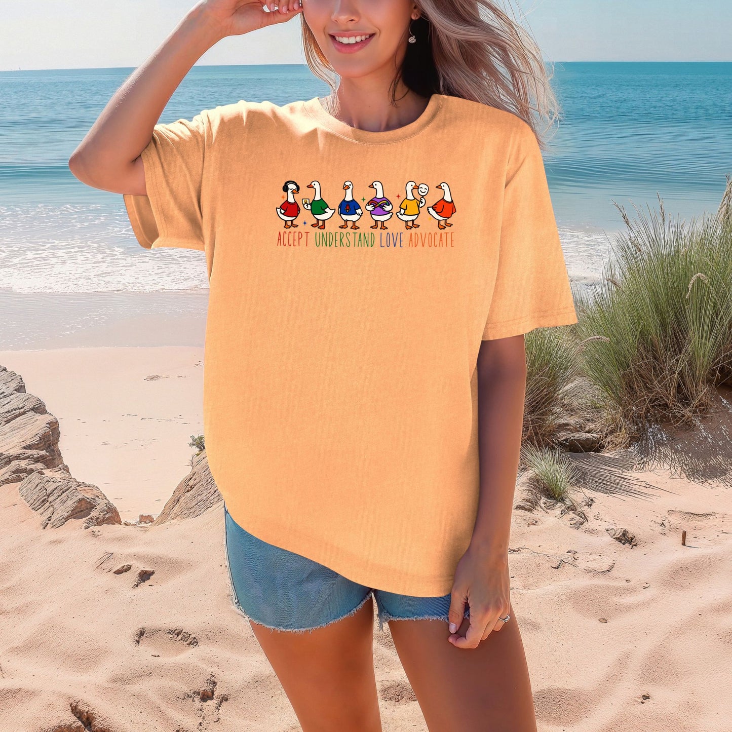 A smiling woman on a sunny beach wearing a warm peachy orange oversized unisex t-shirt. The shirt features six cartoon duck characters lined up wearing colorful outfits. The words Accept Understand Love Advocate are printed below in multicolor lettering with star sparkle accents.