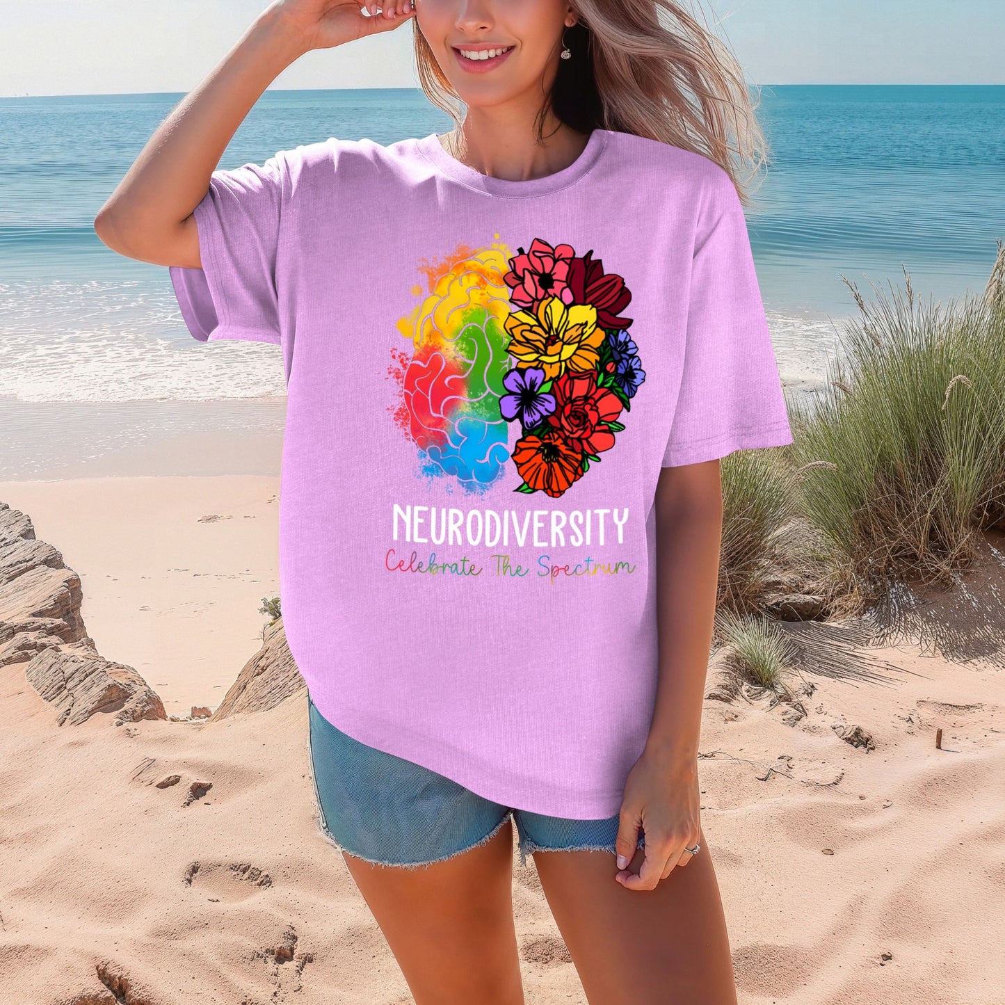 Woman on a beach wearing a soft lavender purple oversized t-shirt with a rainbow floral brain graphic. The left brain features colorful watercolor splashes and the right side blooms into vibrant wildflowers in pink, red, yellow, purple, and orange. Text reads NEURODIVERSITY and Celebrate The Spectrum in multicolor lettering.