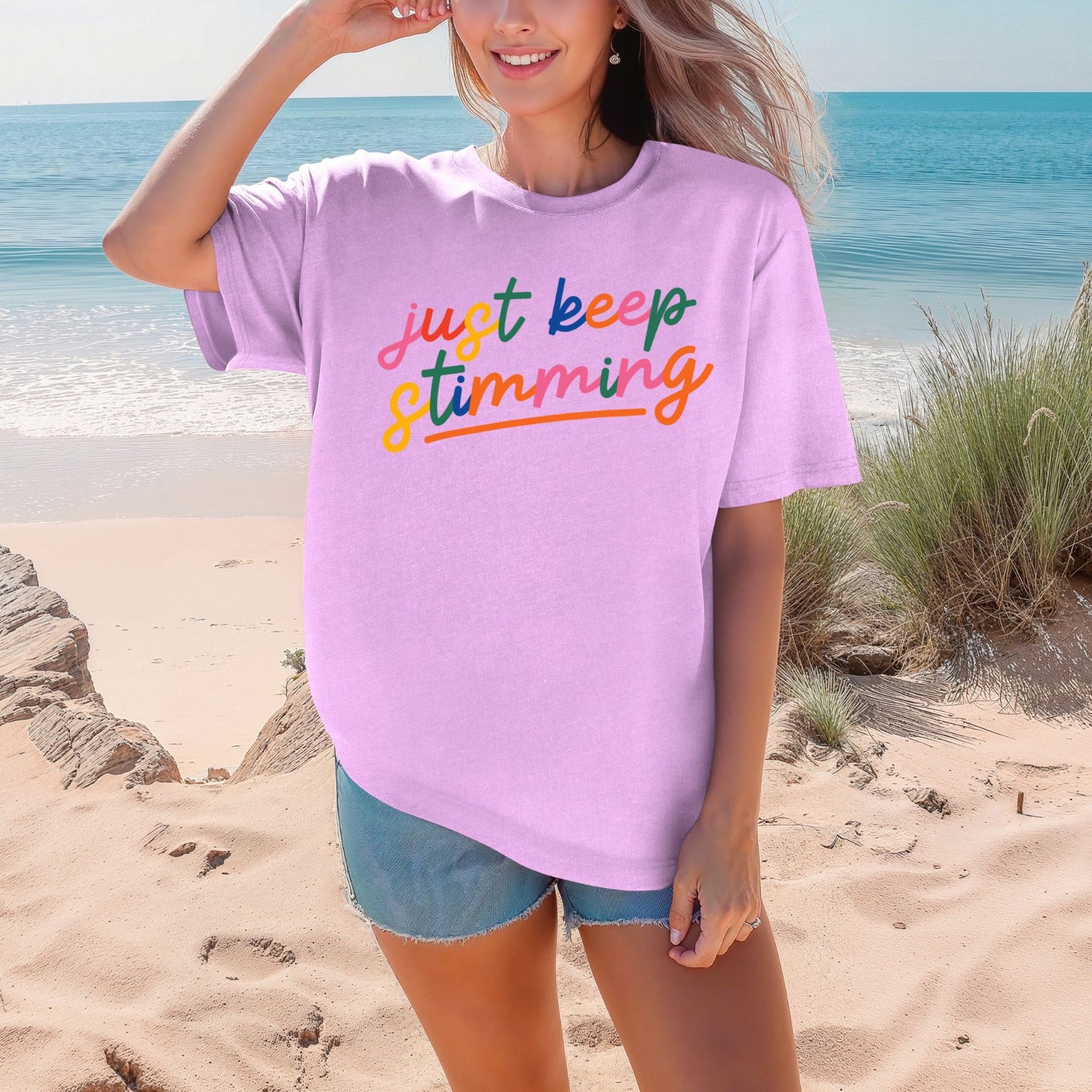 Smiling woman on a beach wearing a soft lavender neon violet oversized unisex t-shirt. The shirt features the phrase just keep stimming in large multicolor rainbow script lettering in pink, blue, green, yellow, and orange with an orange underline beneath the word stimming.