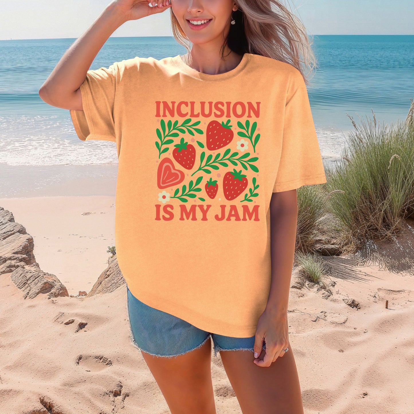 A smiling woman on a sunny beach wearing a warm peachy-orange Neon Cantaloupe color shade, oversized unisex t-shirt. The shirt features a cheerful strawberry botanical graphic with five red strawberries of varying sizes, a peach heart-shaped strawberry, bright green leafy branches, small cream daisy flowers, and soft pink dot accents. The words 'Inclusion Is My Jam' are printed in bold coral red retro lettering above and below the graphic.