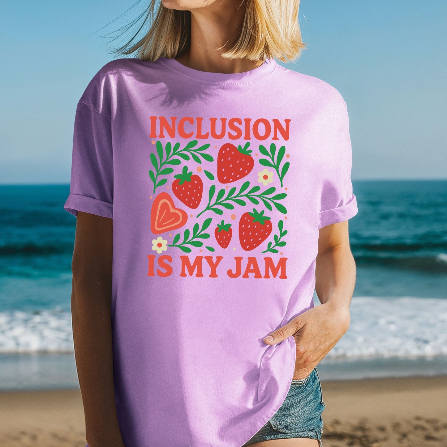 A woman at the beach wearing a soft lavender purple called Neon Violet, oversized unisex t-shirt with rolled sleeves. The shirt displays a vibrant strawberry garden graphic with five red strawberries of varying sizes, a large peach heart-shaped strawberry, lush green leafy branches spreading across the design, small cream daisy flowers with yellow centers, and scattered peach dot accents. The phrase 'Inclusion Is My Jam' is printed in bold coral red retro lettering above and below the graphic.