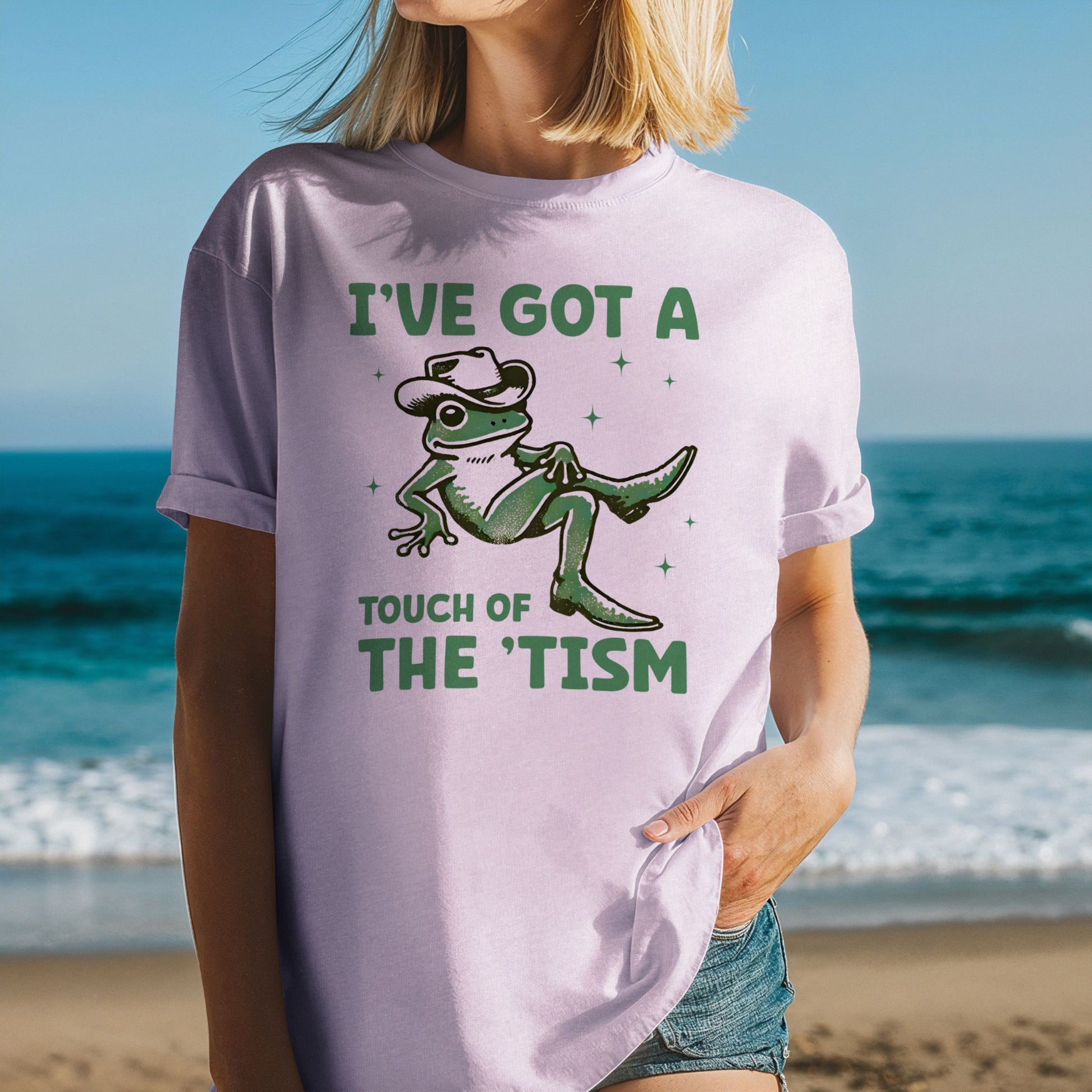 Woman at the beach wearing an Orchid soft lavender Comfort Colors t-shirt with rolled sleeves. The shirt features a green illustrated frog wearing a cowboy hat and cowboy boots with sparkle accents. Text above reads I've Got a and text below reads Touch of The Tism in bold green lettering.