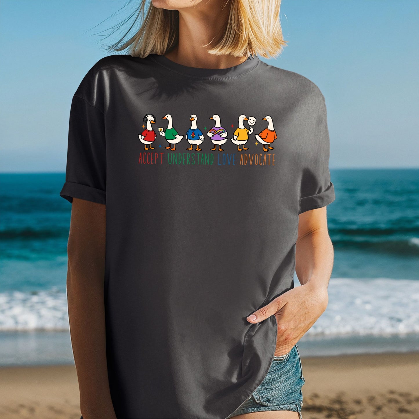 A woman at the beach wearing a dark charcoal gray oversized unisex t-shirt with rolled sleeves. The shirt features six cartoon duck characters each wearing a different colorful outfit. The words Accept Understand Love Advocate are printed below the ducks in multicolor lettering with sparkle details.