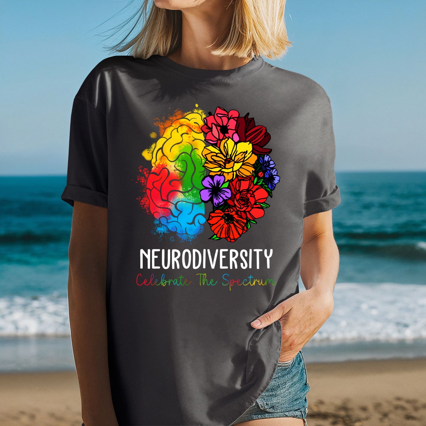 Woman at the beach wearing a dark charcoal gray oversized t-shirt with rolled sleeves and a vivid rainbow floral brain graphic. The left brain has orange, yellow, green, red, and blue watercolor splashes. The right side bursts into pink, red, yellow, purple, and orange wildflowers. Text reads NEURODIVERSITY and Celebrate The Spectrum in multicolor lettering.