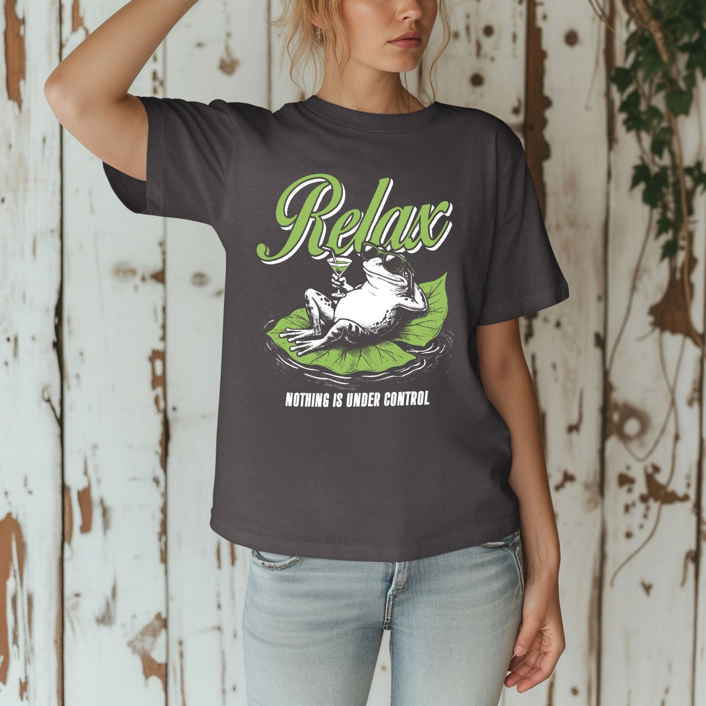 Woman against a rustic white wooden wall wearing a Pepper dark charcoal gray Comfort Colors unisex t-shirt. The shirt features a retro illustrated frog wearing sunglasses, lounging on a green lily pad holding a cocktail glass. The word Relax appears in bold green script above and Nothing Is Under Control in white block letters below.