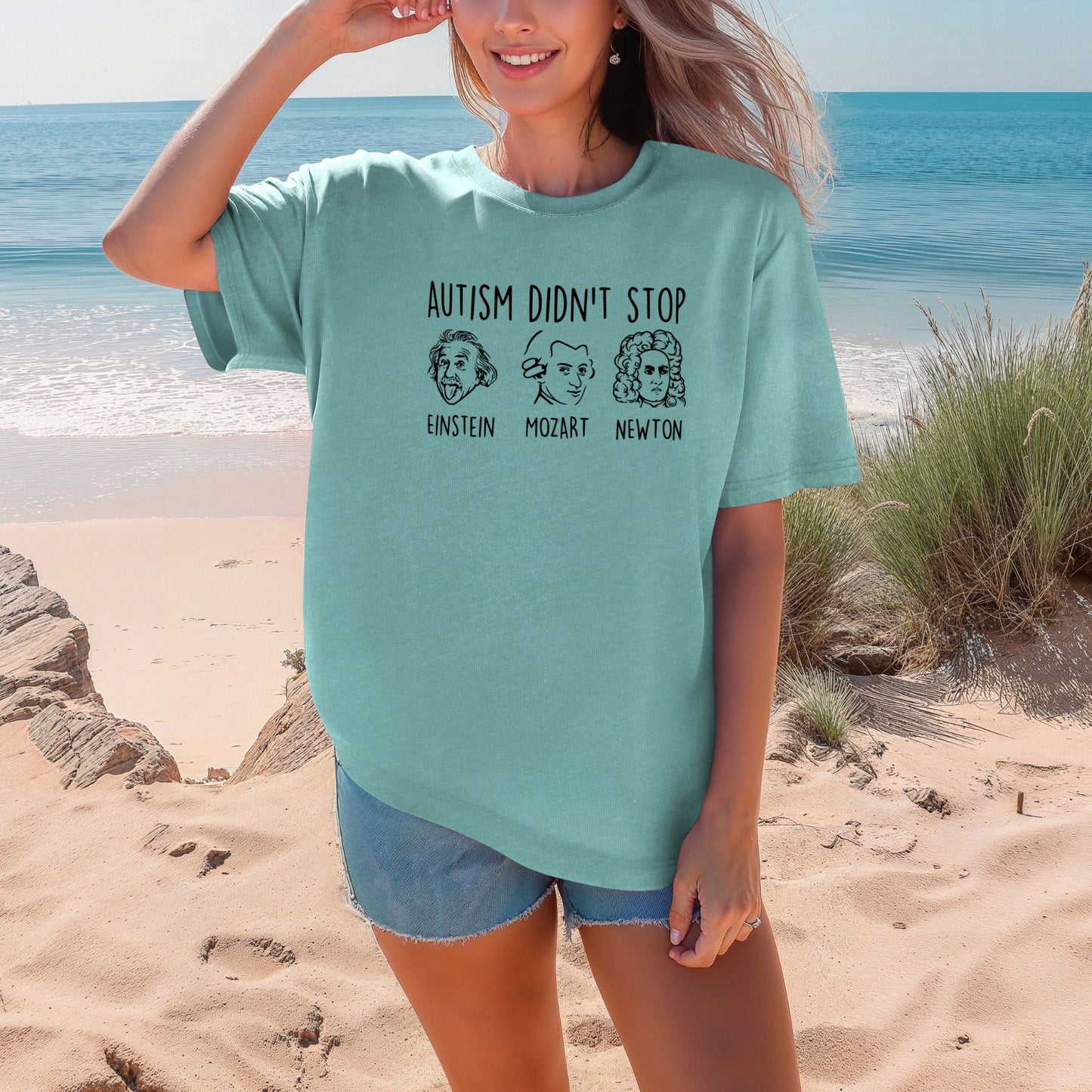 Smiling woman on a beach wearing a Sea Foam mint teal Comfort Colors oversized t-shirt. The shirt features hand-drawn style portraits of Einstein, Mozart, and Newton in black ink with the text Autism Didn't Stop printed above in bold black lettering and each name labeled below their portrait.