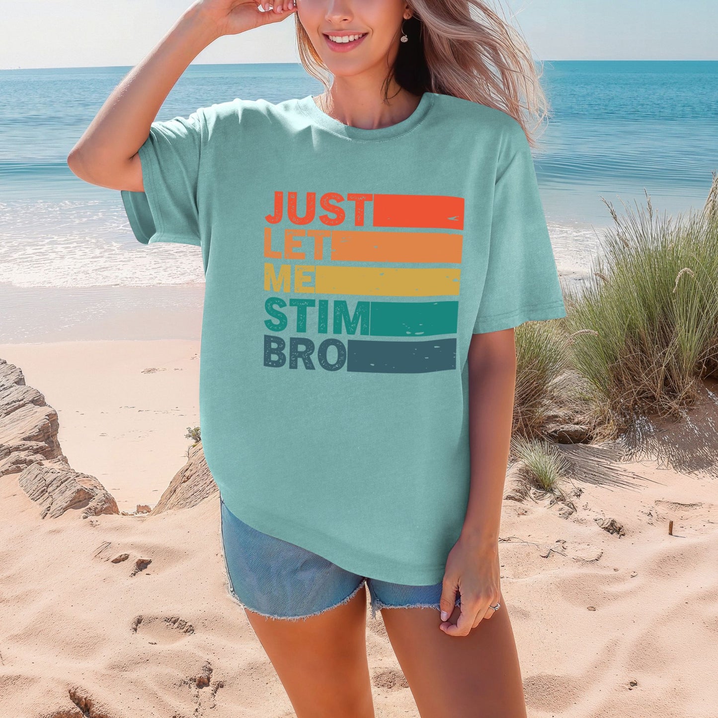 Smiling woman on a beach wearing a Sea Foam mint teal Comfort Colors oversized t-shirt. The shirt features bold retro stripe lettering reading Just Let Me Stim Bro stacked vertically with each word on a colored stripe in orange, peach, yellow, teal, and dark teal with a distressed vintage texture.
