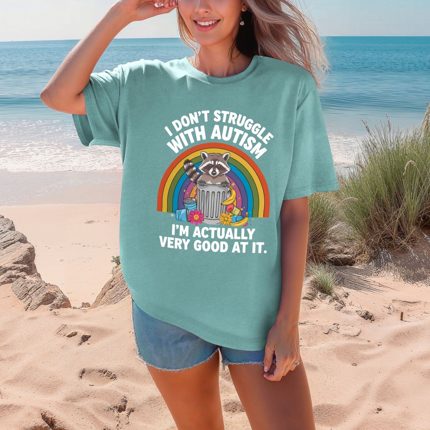 Comfort Colors 9018 youth t-shirt in Sea Foam minty teal. A woman stands on a sandy beach with dunes and ocean behind her. The shirt features a raccoon in a trash can in front of a rainbow with the text I Don't Struggle With Autism I'm Actually Very Good At It.