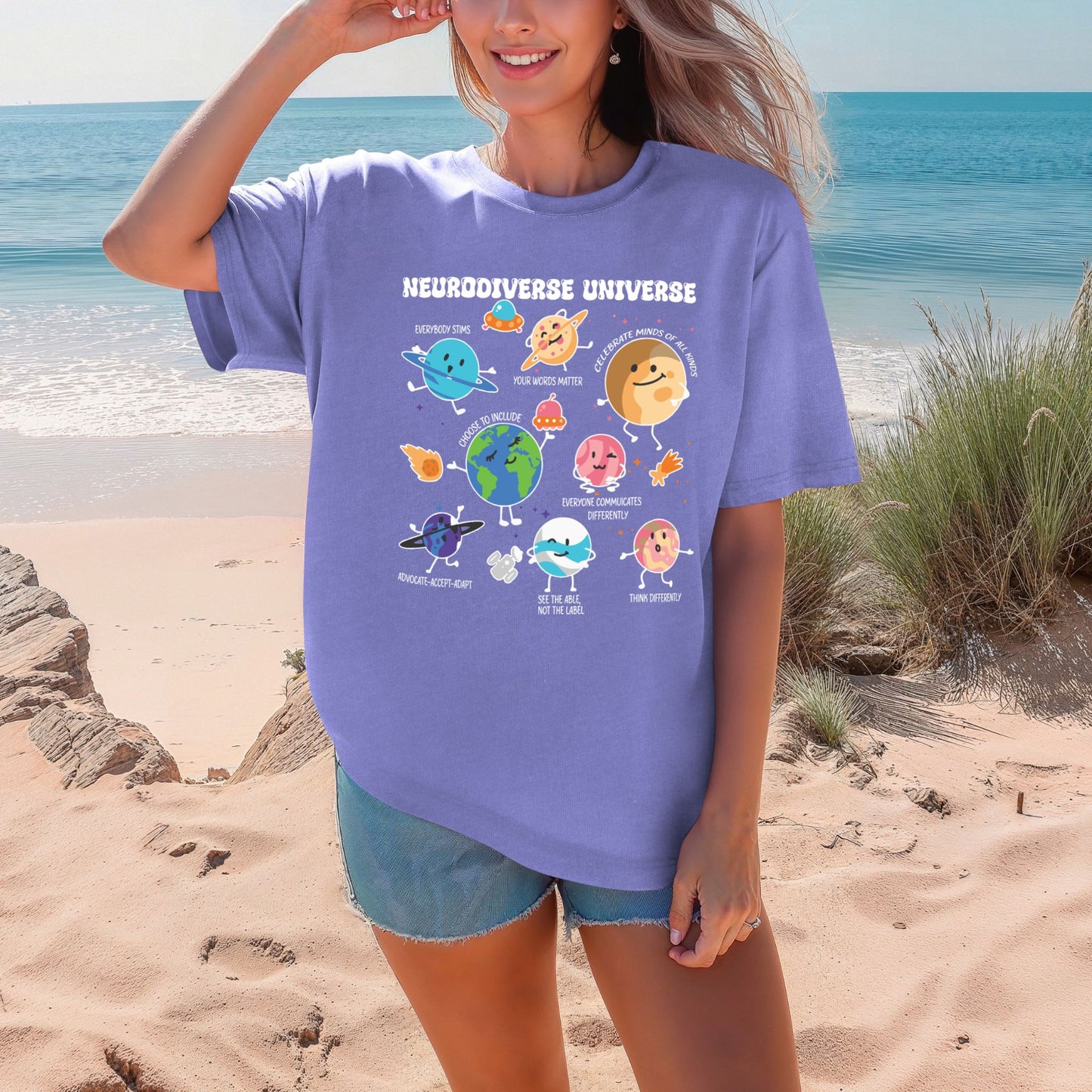Smiling woman on a beach wearing a Violet purple Comfort Colors oversized t-shirt. The shirt features colorful cartoon planet characters each labeled with an empowering neurodiversity message. The title Neurodiverse Universe is printed at the top in bold white lettering.