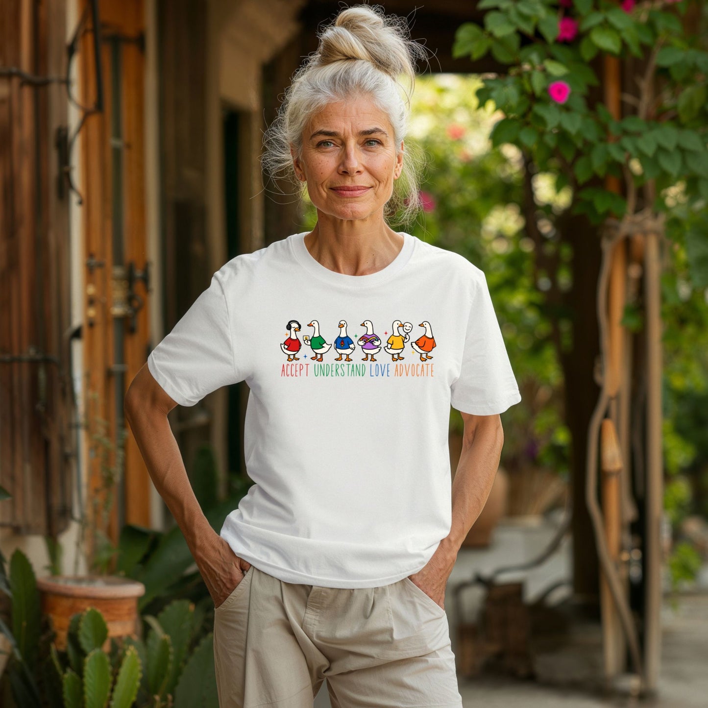 An older woman with white hair standing in a garden courtyard wearing a crisp white unisex t-shirt. The shirt features six cheerful cartoon duck characters lined up in a row wearing colorful outfits. Below the ducks the text reads Accept Understand Love Advocate in bright rainbow colored lettering.