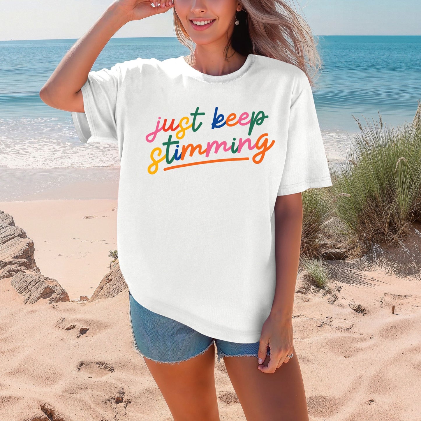Smiling woman on a beach wearing a crisp white oversized unisex t-shirt. The shirt features the phrase just keep stimming in large multicolor rainbow script lettering in pink, blue, green, yellow, and orange with an orange underline beneath the word stimming.