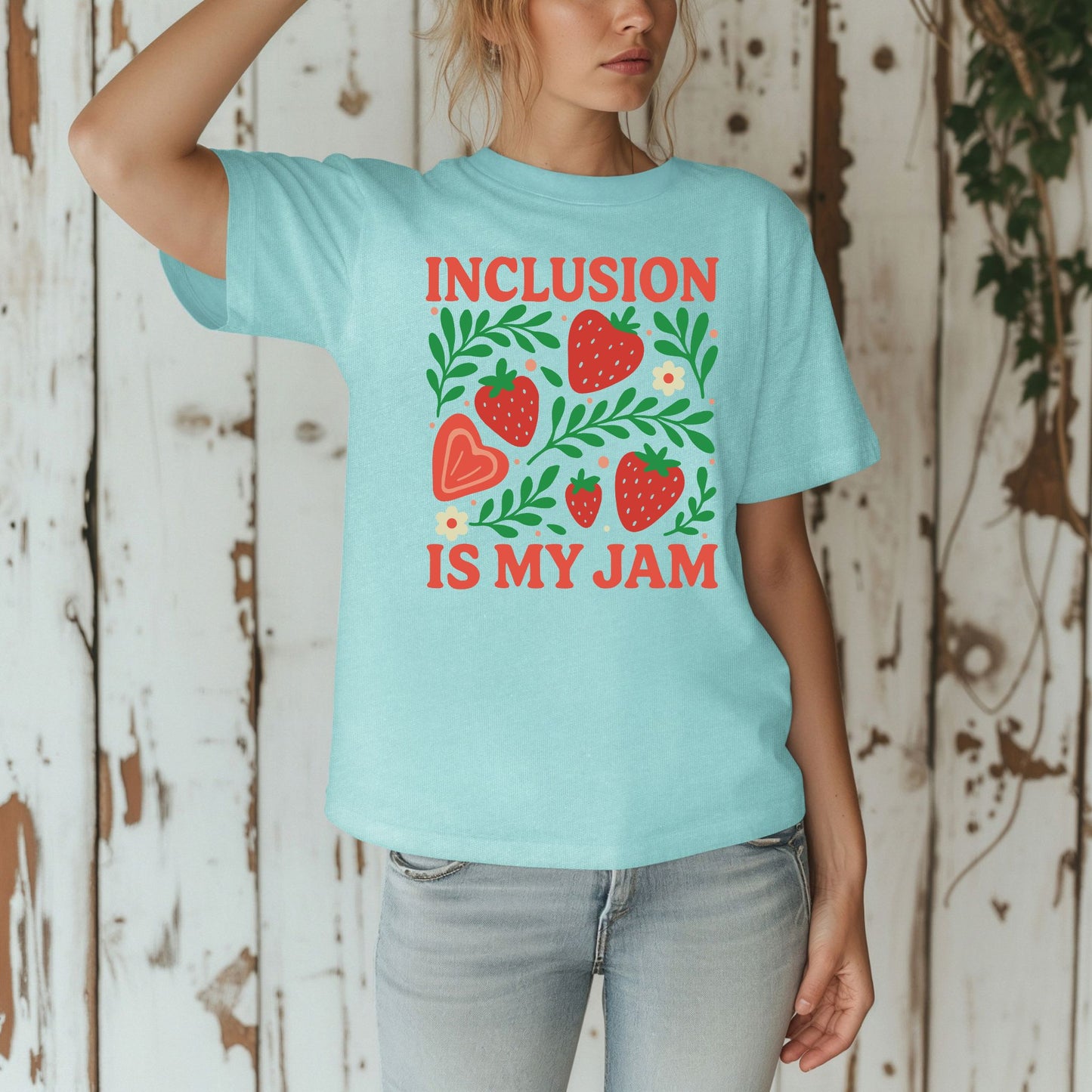 A woman standing against a rustic white wooden wall wearing a chalky mint blue oversized unisex t-shirt. The shirt features a fresh strawberry botanical graphic with five red strawberries of varying sizes, a peach heart-shaped strawberry, vibrant green leafy branches, small cream daisy flowers, and peach dot accents. The words 'Inclusion Is My Jam' are printed in bold coral red retro lettering above and below the graphic.