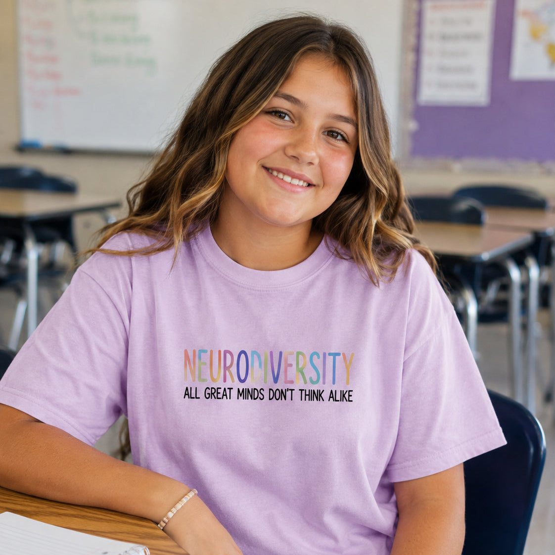 A young girl with long brown hair sits at a desk in a classroom, wearing a light purple t-shirt with the text 'NEURODIVERSITY ALL GREAT MINDS DON'T THINK ALIKE' printed on the front.