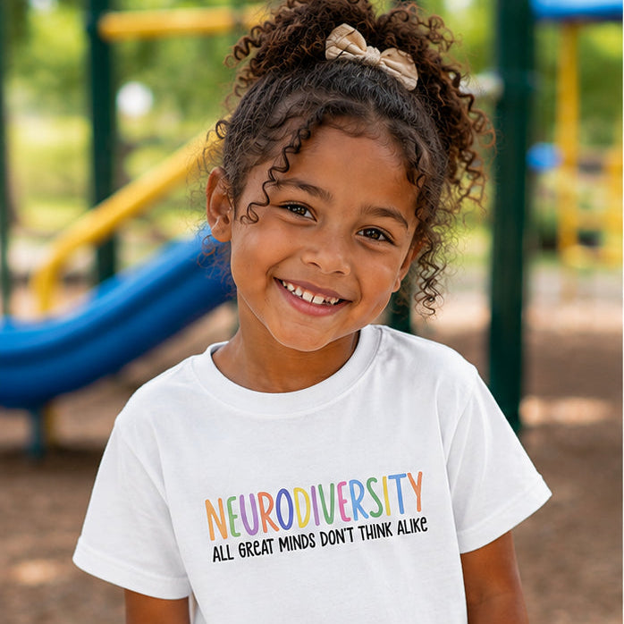 A young girl with curly hair stands on a playground wearing a white graphic t-shirt that reads 'NEURODIVERSITY ALL GREAT MINDS DON'T THINK ALONE' in rainbow-colored lettering, paired with light blue jeans.